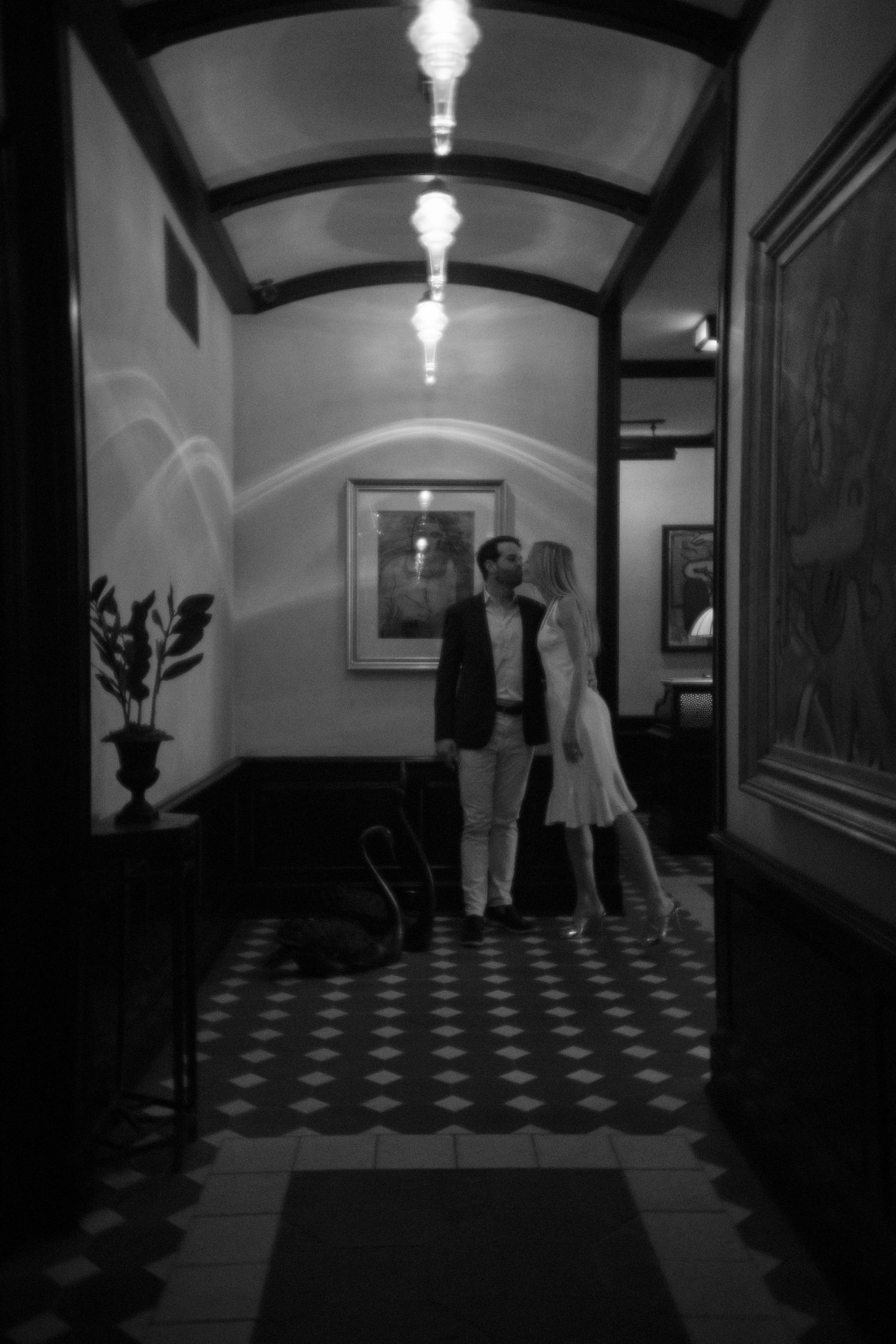 Black and white photo of a hallway with checkered floor and arched ceiling with vintage lighting fixtures.