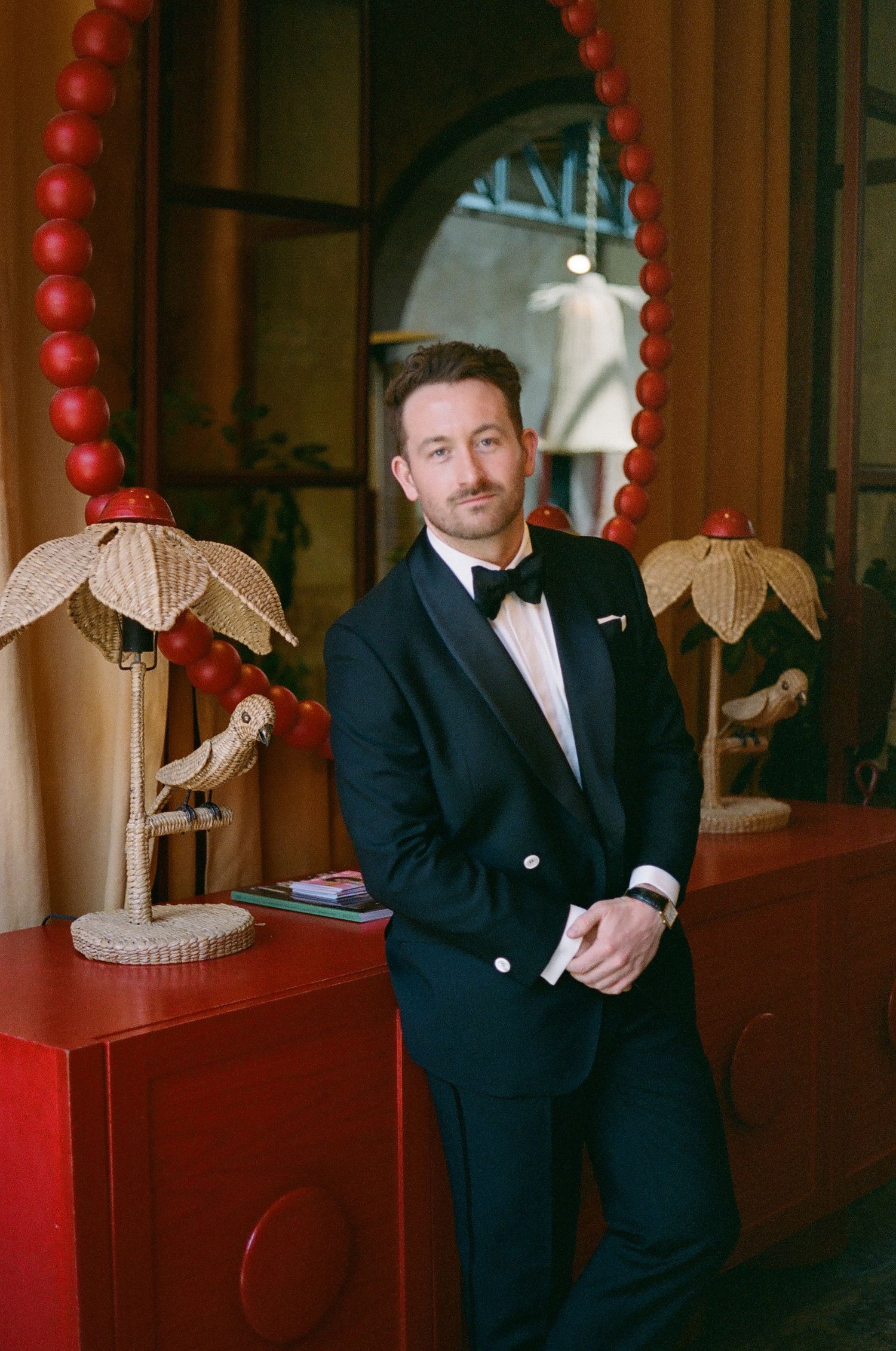 A person in a classic black tuxedo and bow tie stands elegantly next to decorative red beads and floral lamp fixtures. San Miguel Mexico