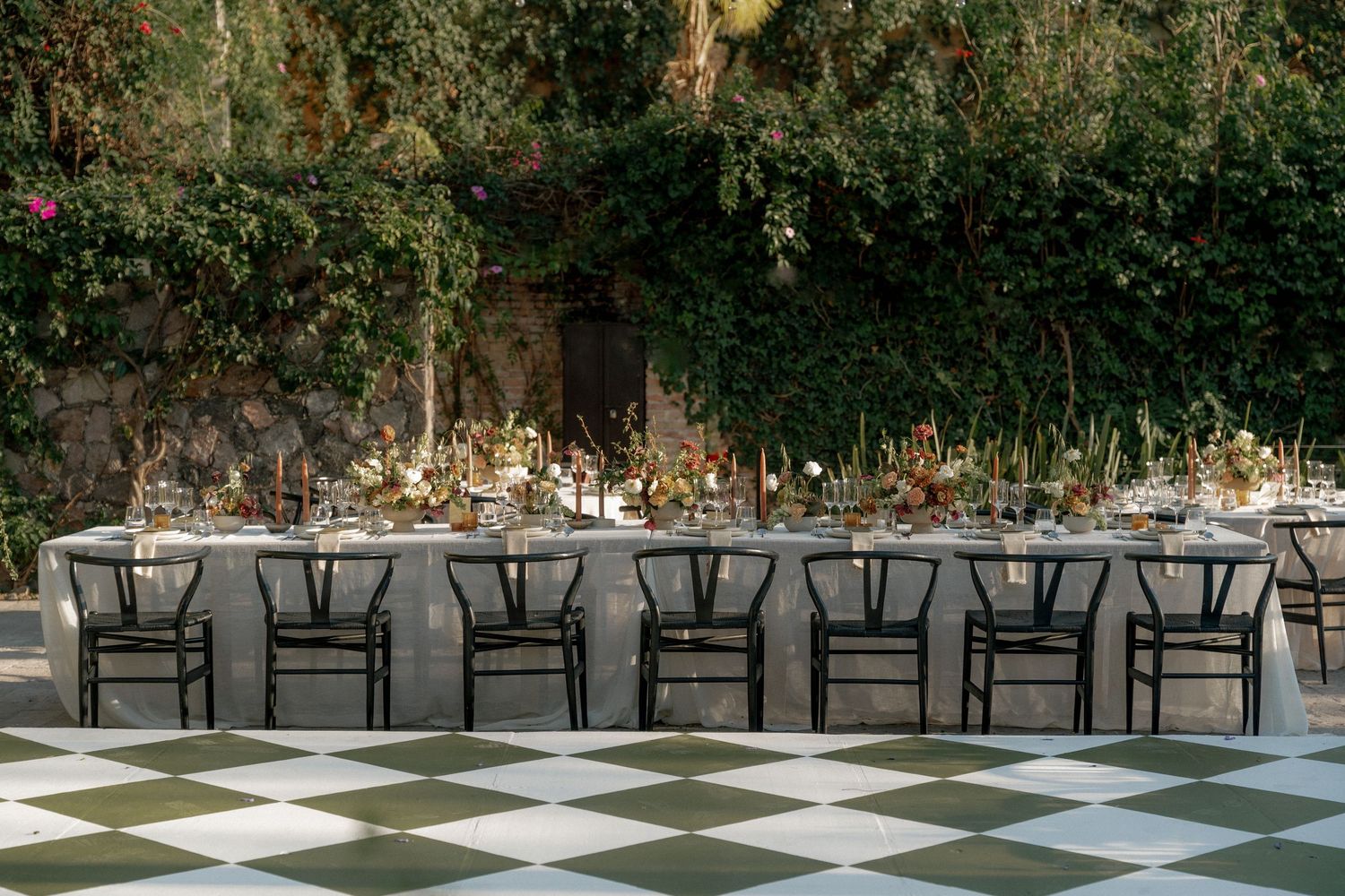 Long reception table with black chairs set against ivy-covered wall on checkered floor.