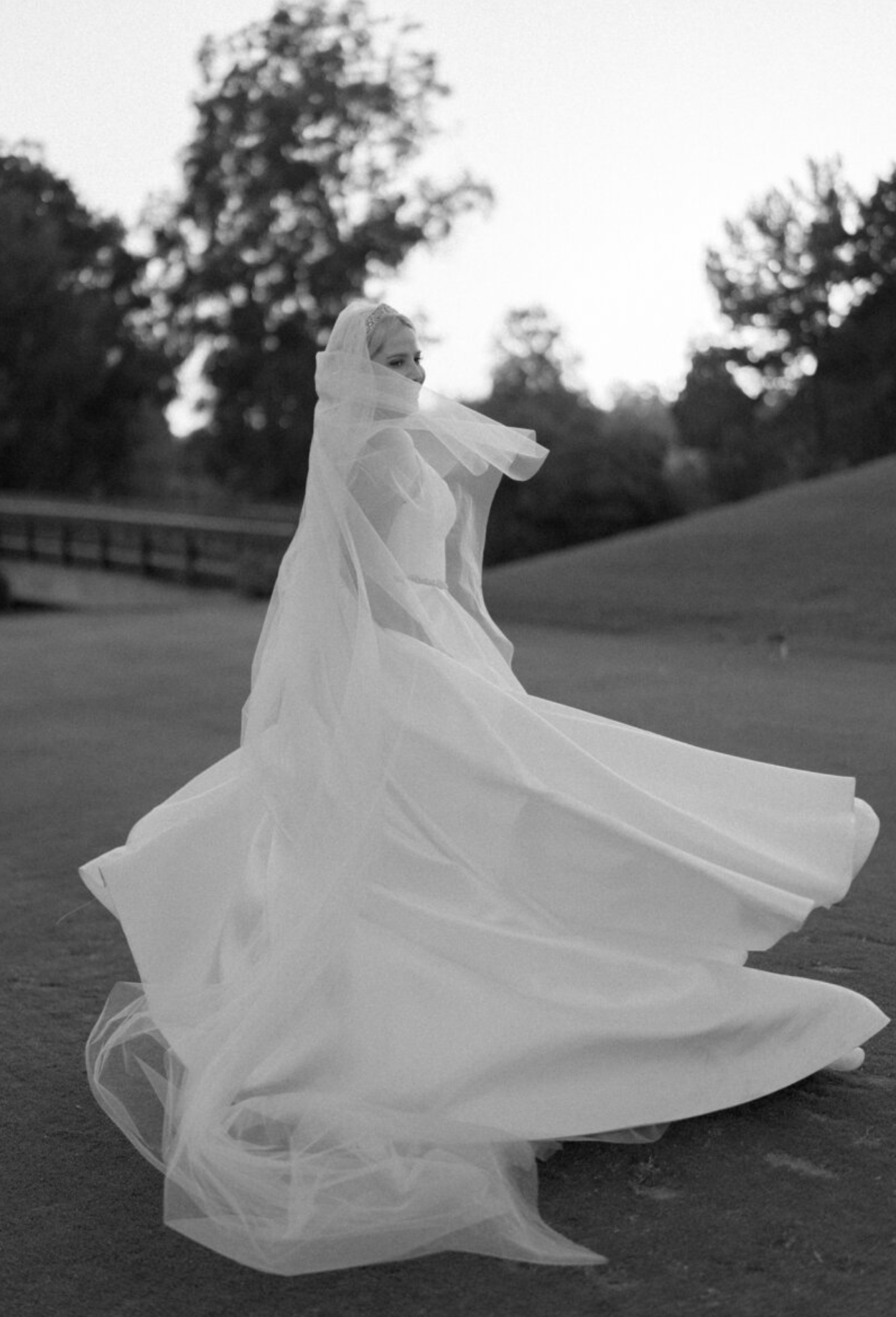 Flowing white wedding dress billowing dramatically in the wind on a grassy field at sunset.