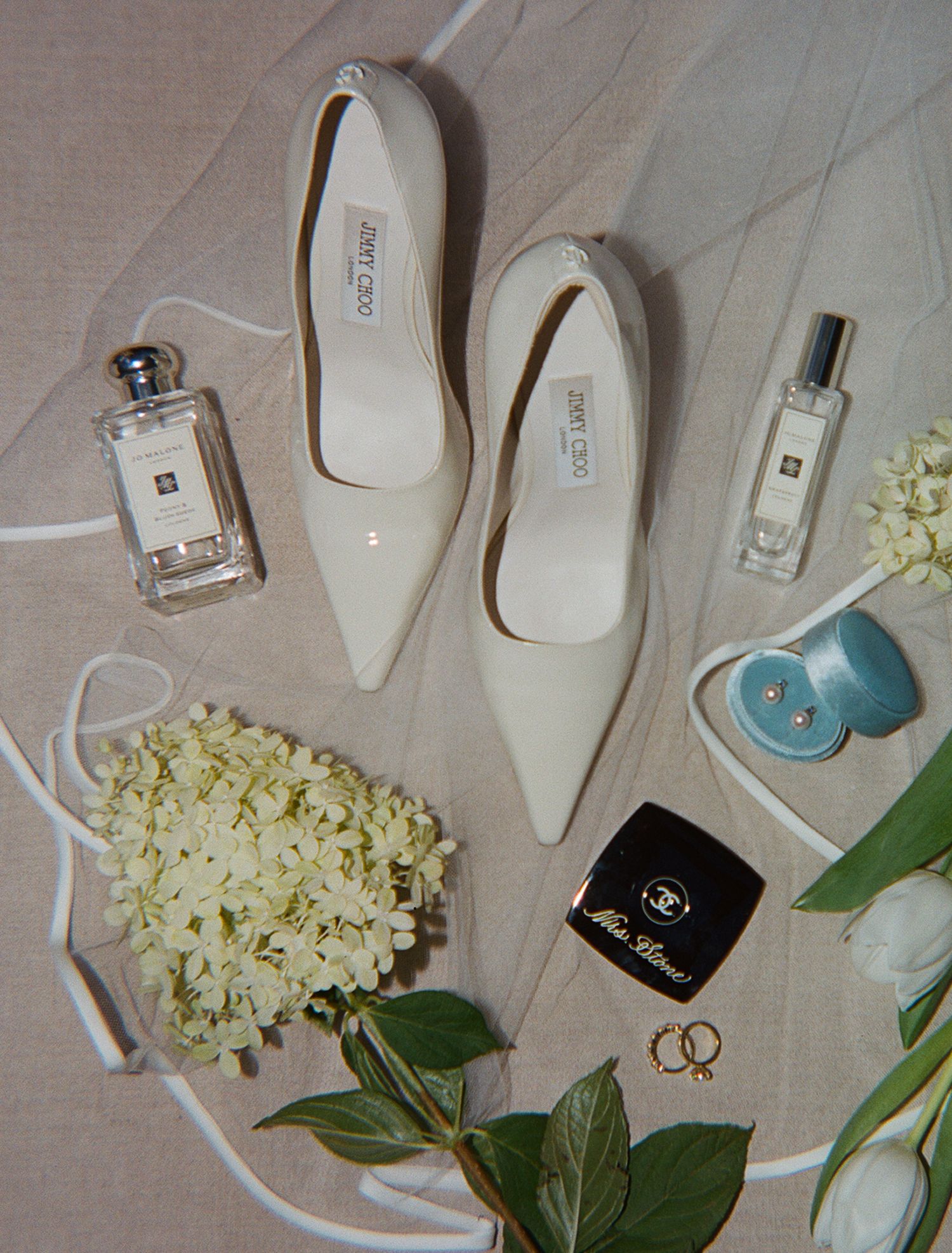 Wedding accessories including white heels, perfume bottles, rings and hydrangea flowers on light fabric.