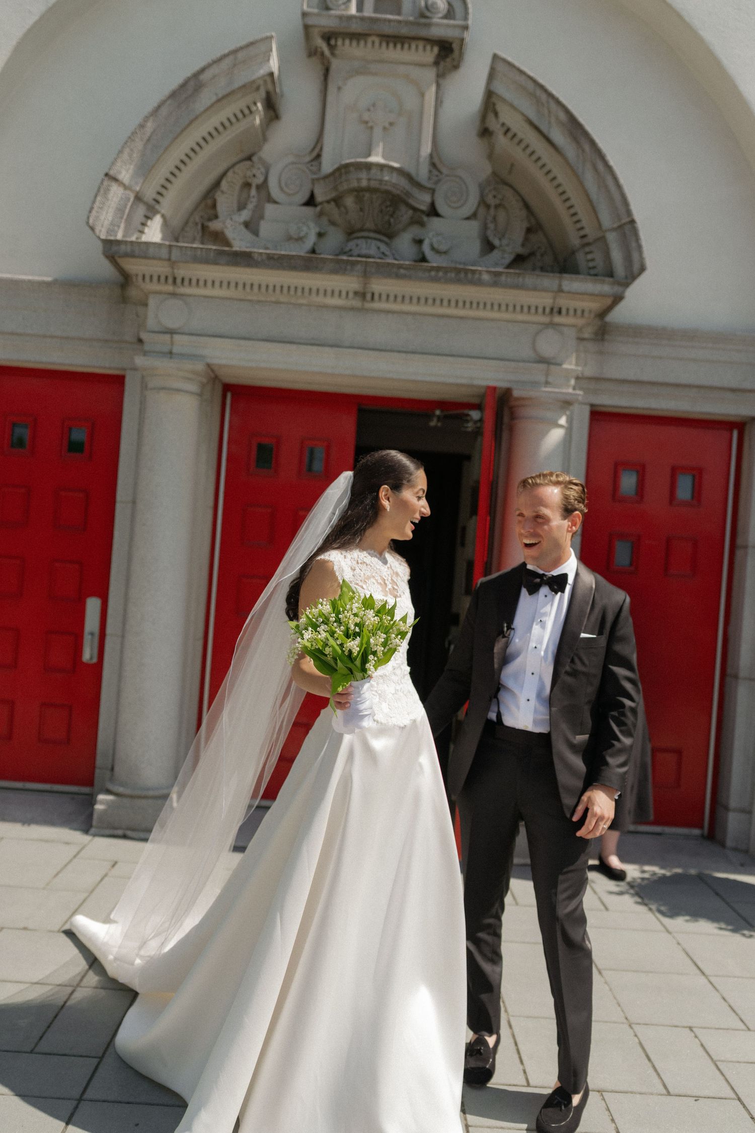 Bride in white gown holding bouquet and groom in black tuxedo walking outside a church with red doors.