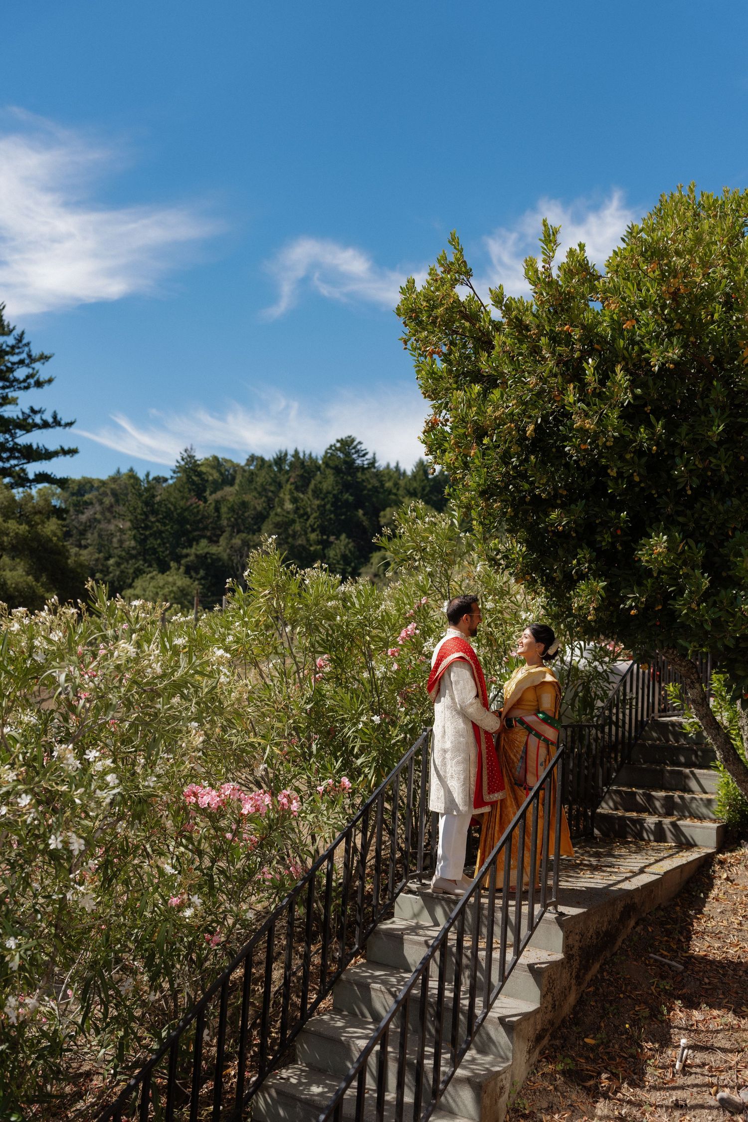 Couple in traditional Indian attire standing on garden steps surrounded by lush greenery and pink flowers under a blue sky.