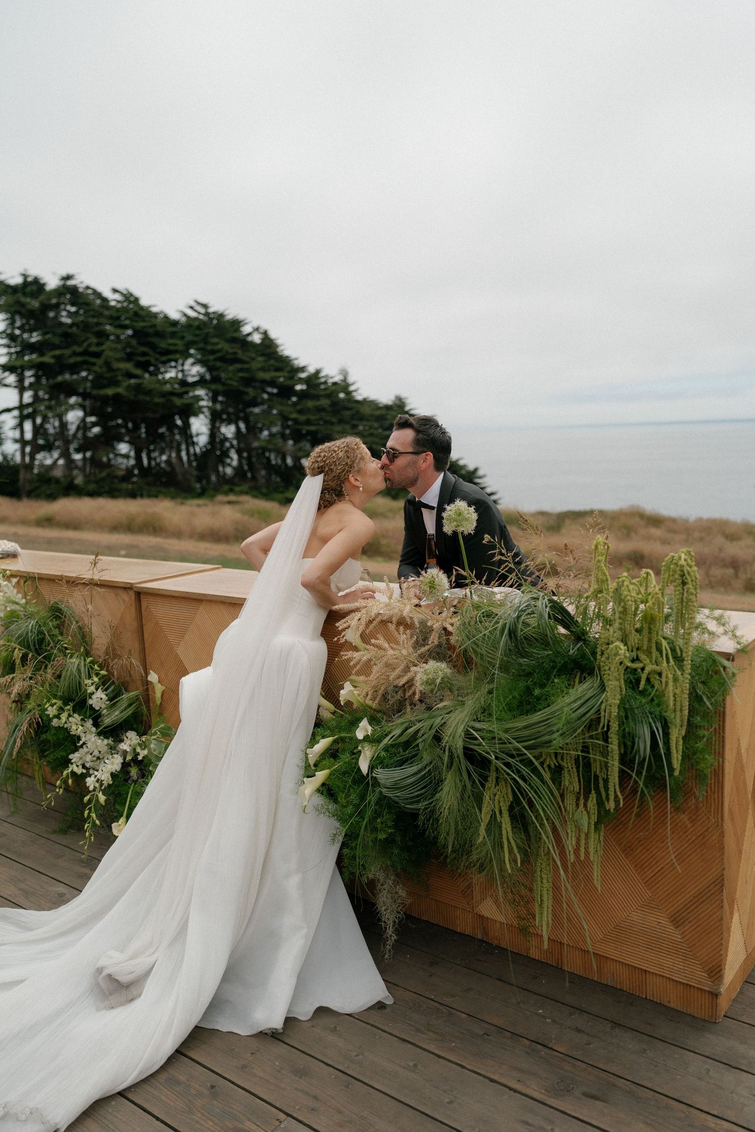 Couple shares intimate moment on wooden deck decorated with greenery and floral arrangements.