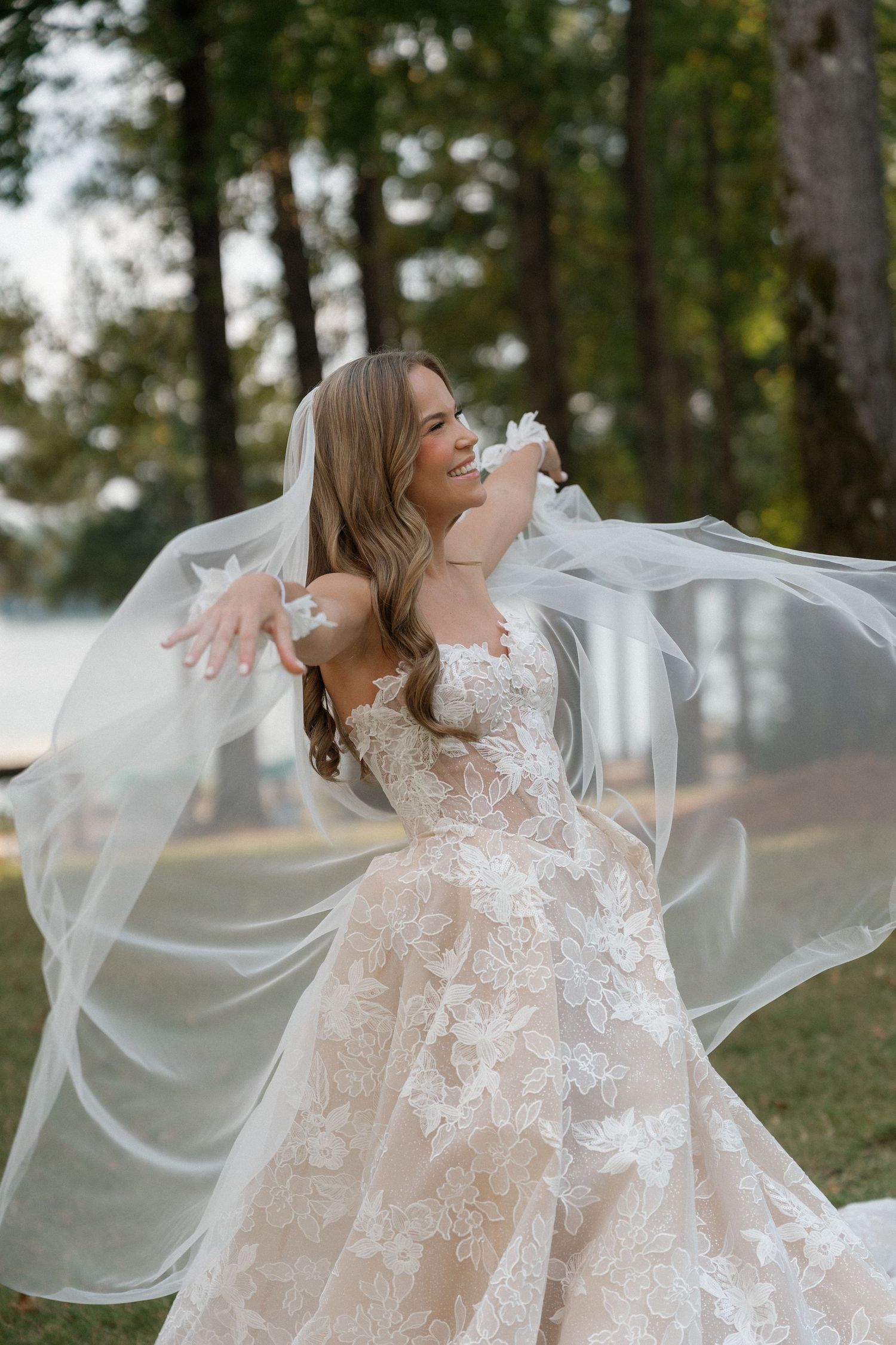 A bride in a lace wedding gown twirls joyfully outdoors with her veil flowing in the breeze.