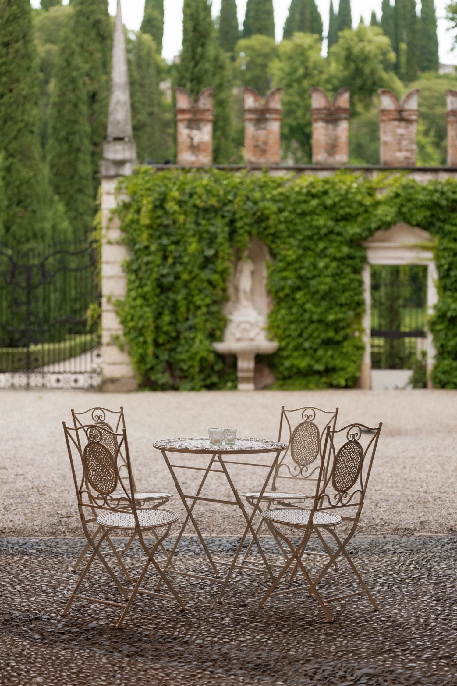 Vintage wrought iron bistro table and chairs set on cobblestone in a lush Italian garden with ivy walls.