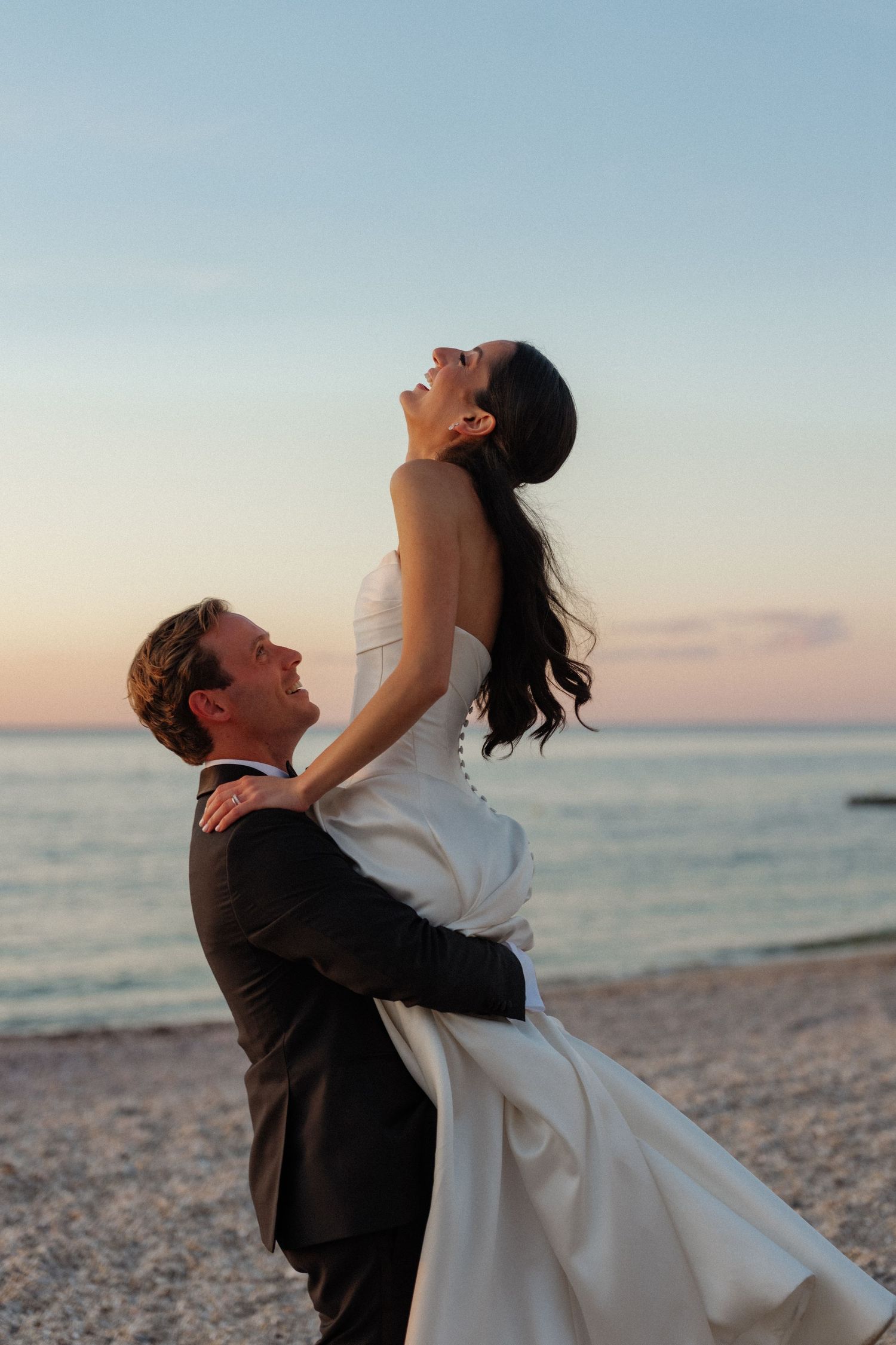 Groom lifts joyful bride on a beach at sunset with ocean backdrop during wedding photo session.
