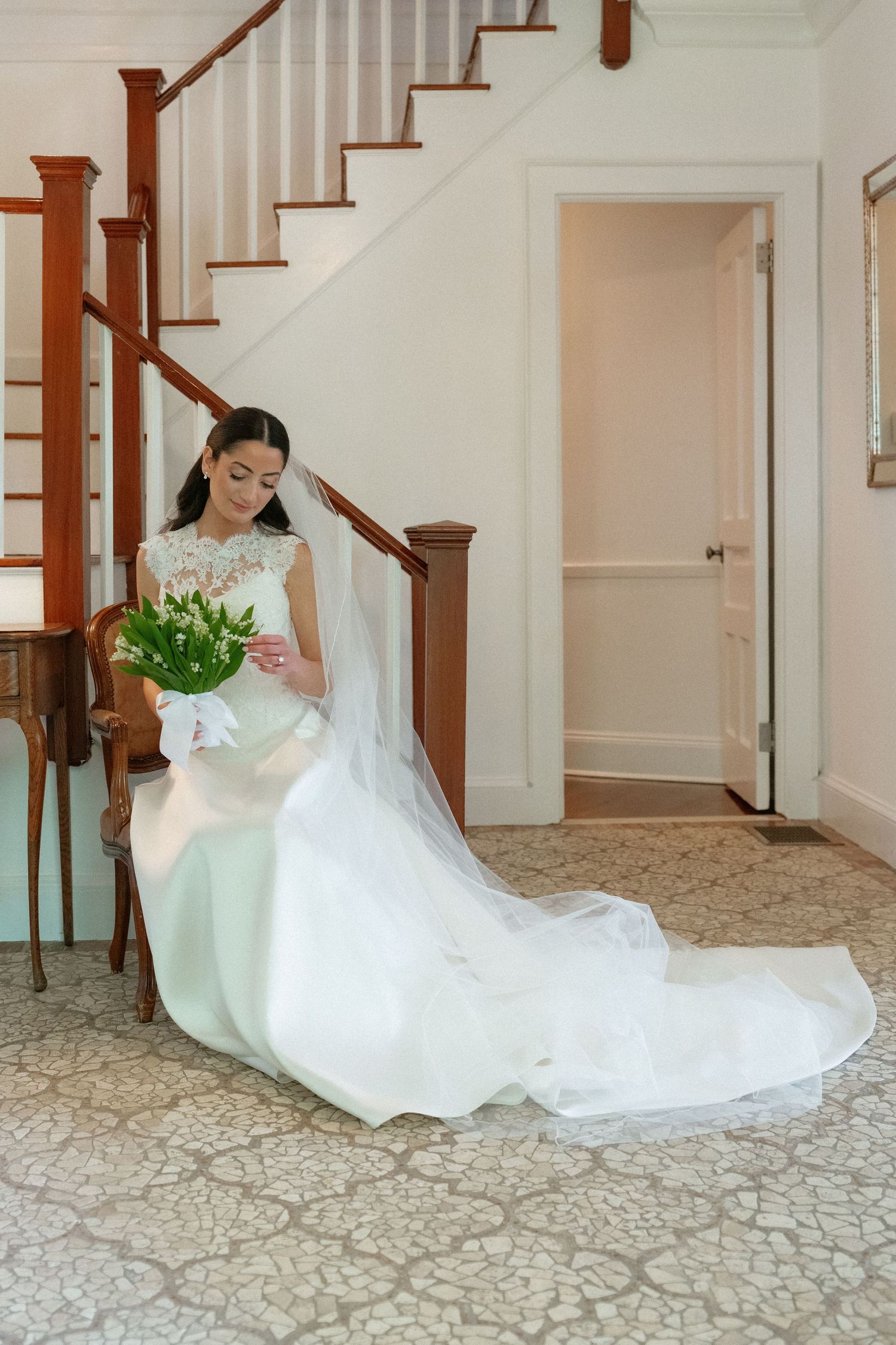 Bride in lace wedding gown with long veil sitting gracefully on chair holding white tulip bouquet.