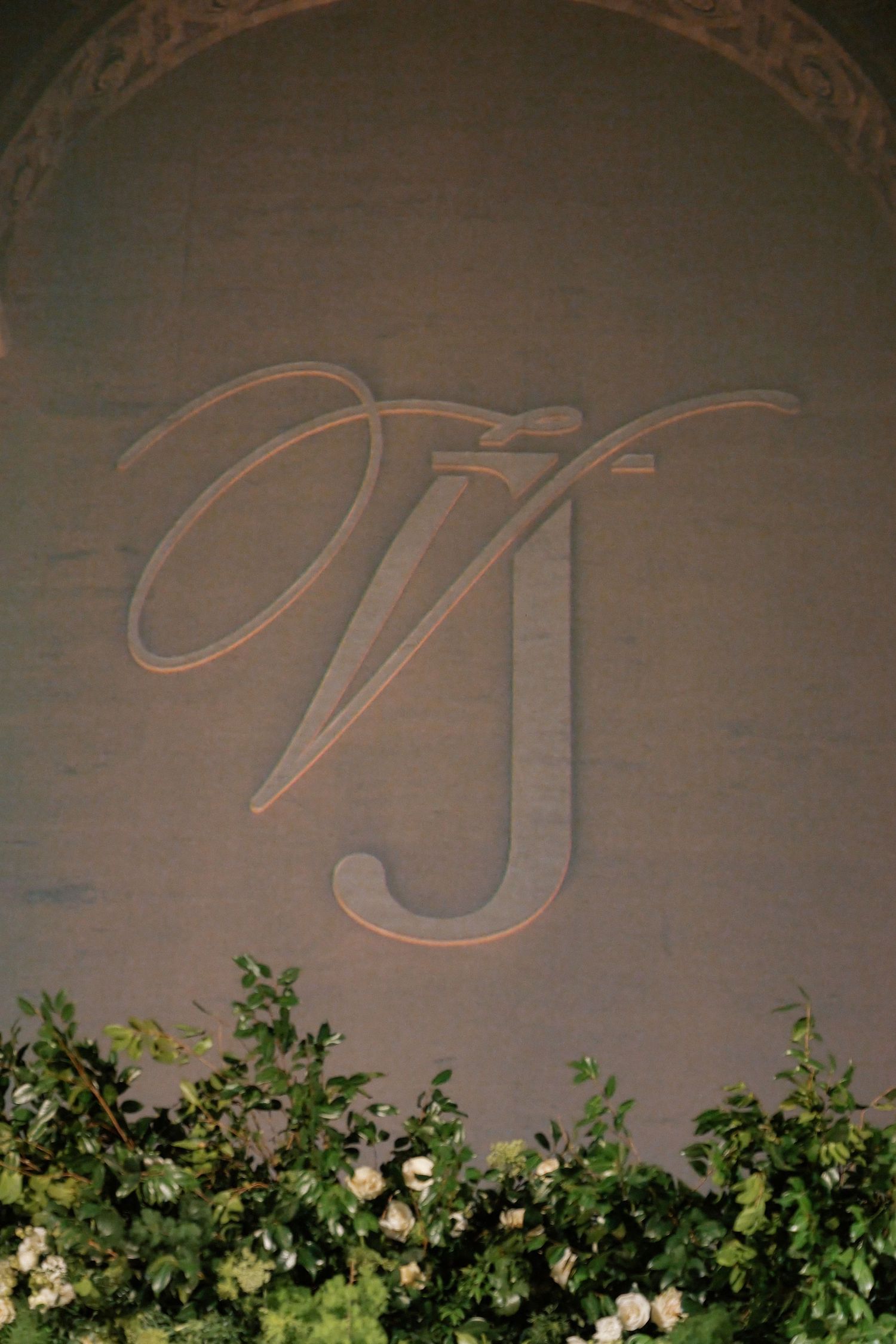 An ornate monogram 'W' carved into a stone wall, surrounded by greenery.