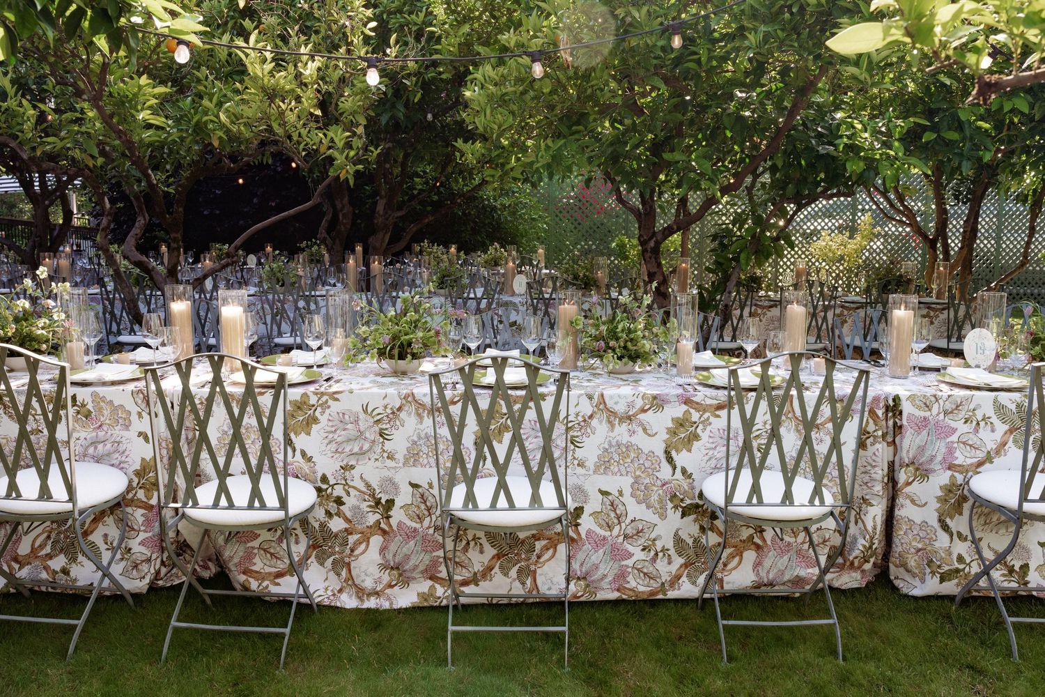 Elegant outdoor garden dining table with floral tablecloth, lattice chairs, candles, and lush greenery for a wedding reception.
