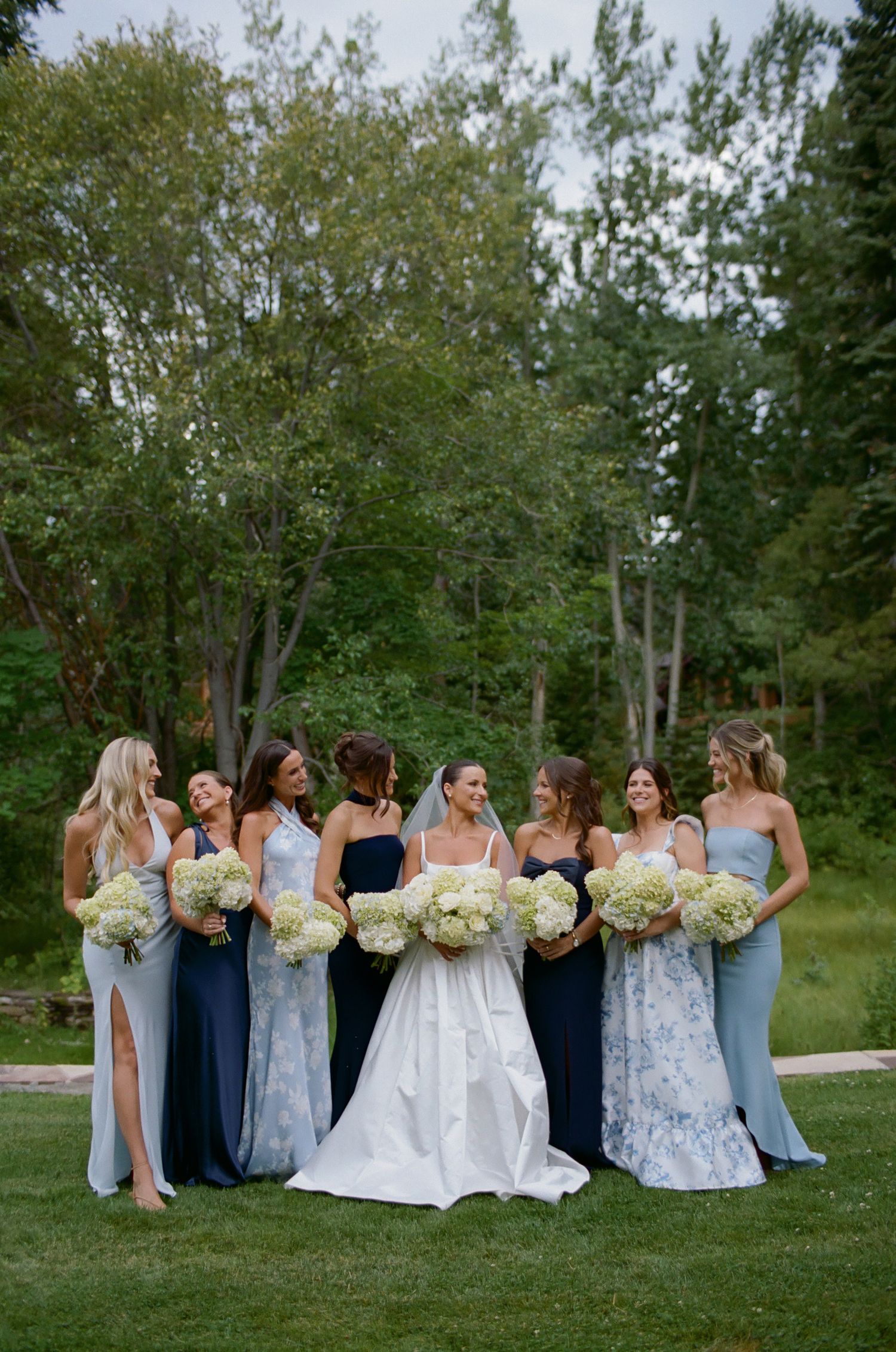 Wedding party in mixed blue and floral dresses poses together on a grassy lawn with bouquets.