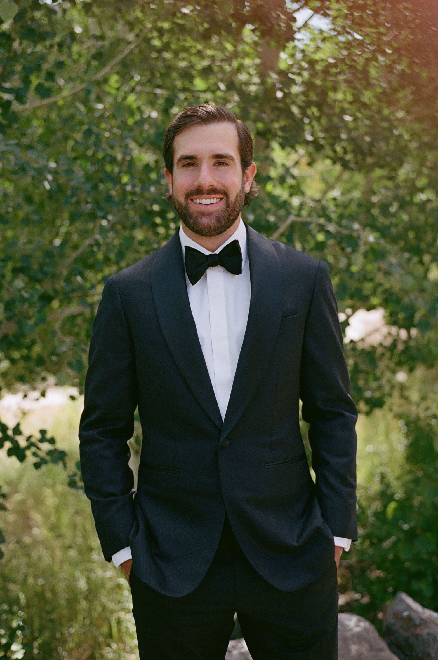 Person wearing black tuxedo and bowtie standing against leafy green background.