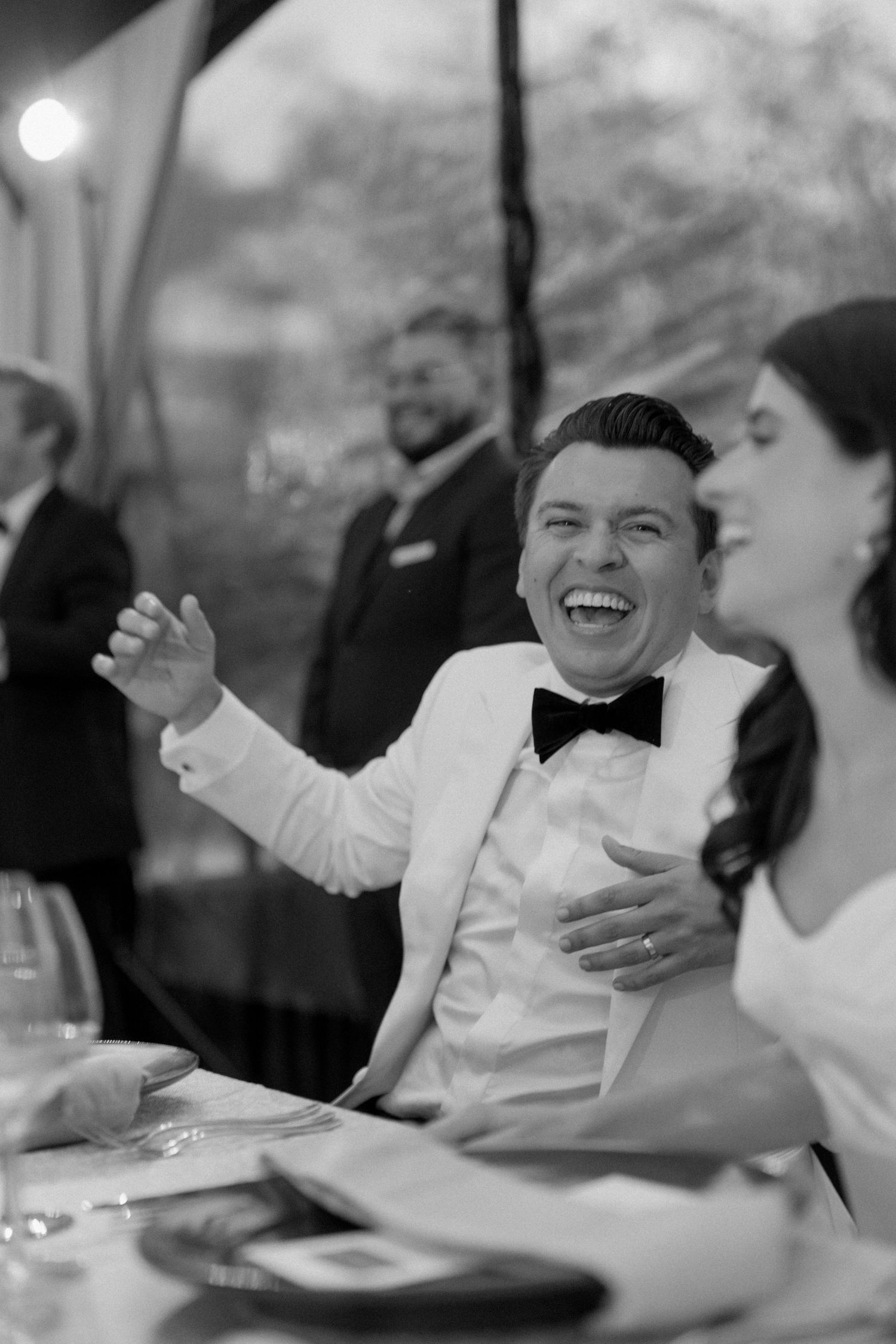 Wedding guests share laughter during a reception dinner under evening lighting.
