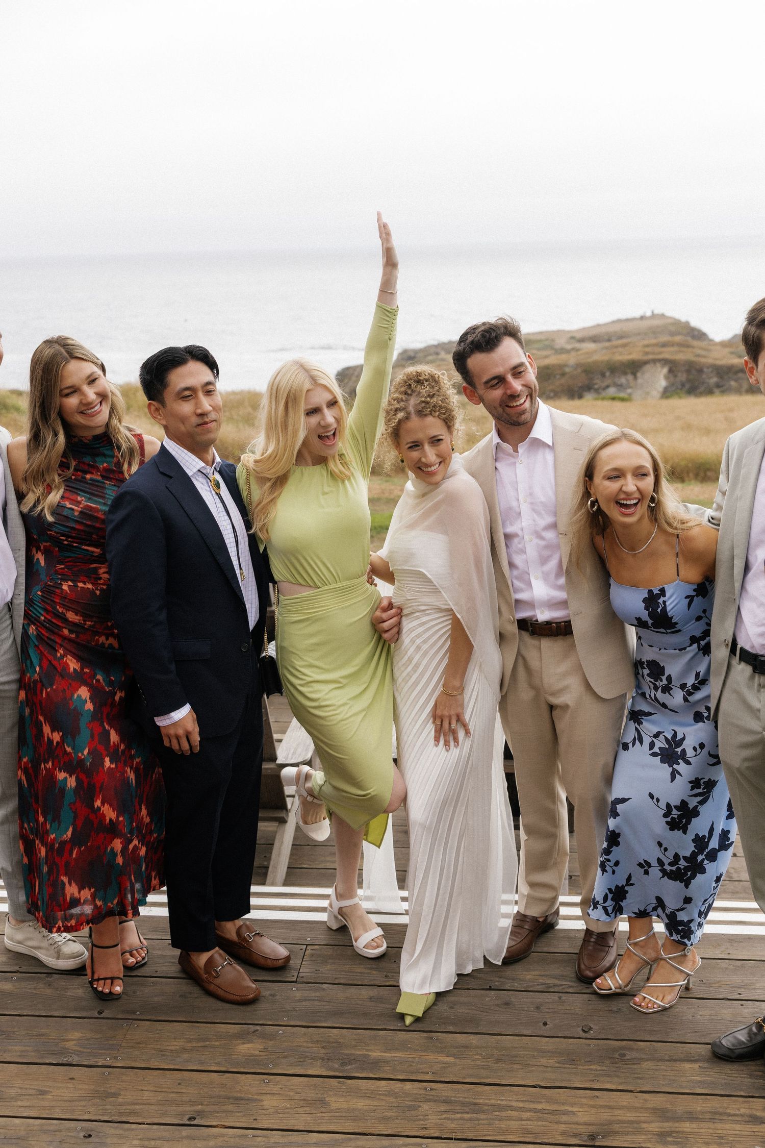 Wedding party posing playfully on wooden deck with coastal background.