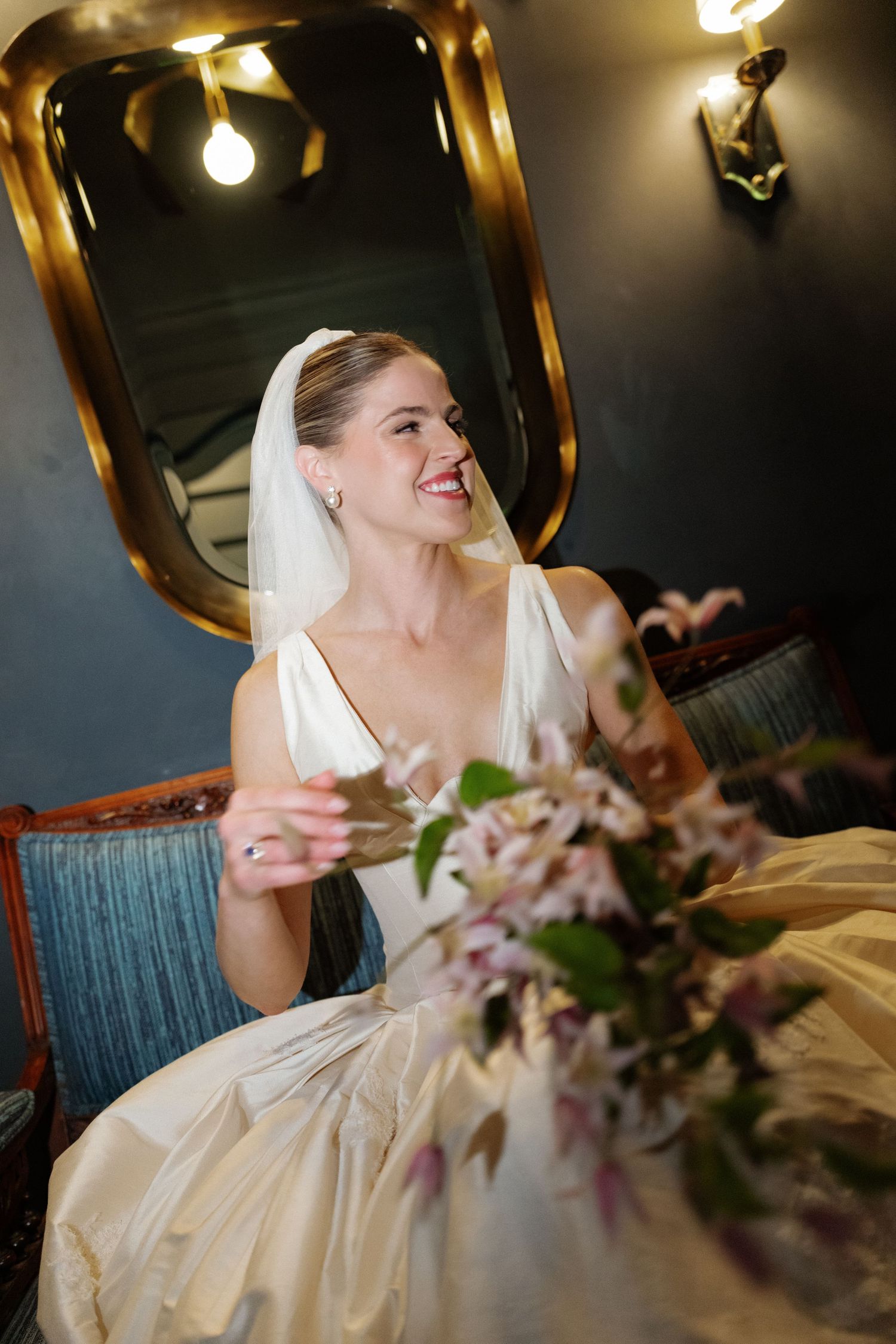 Smiling bride in white gown and veil seated indoors near gold mirror holding a teacup.
