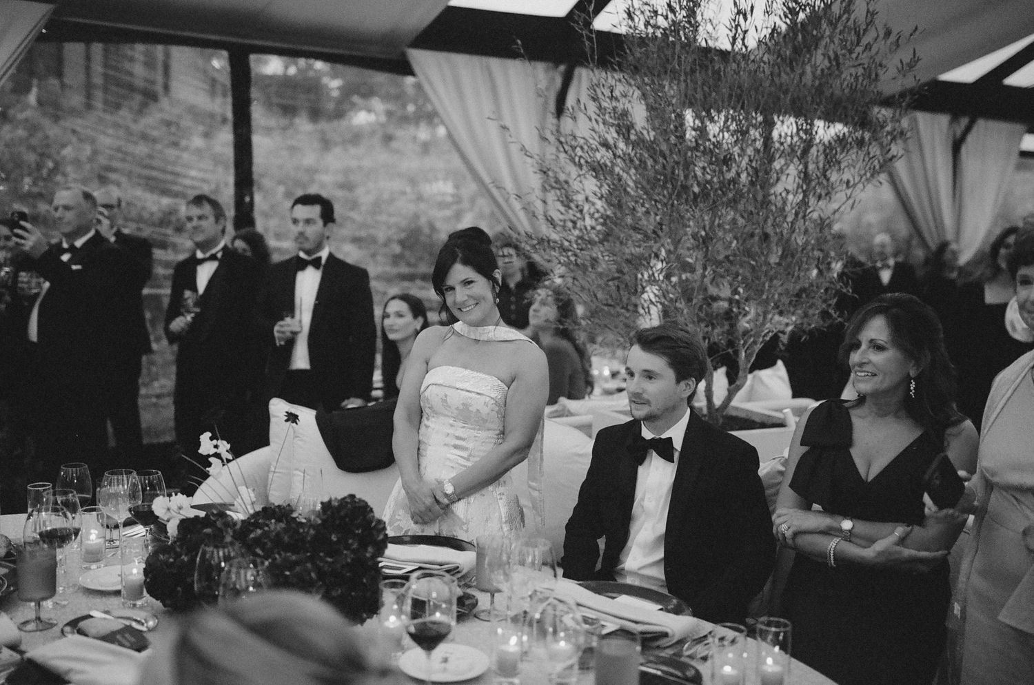 Wedding reception guests enjoying a formal dinner celebration under elegant white tent lighting.
