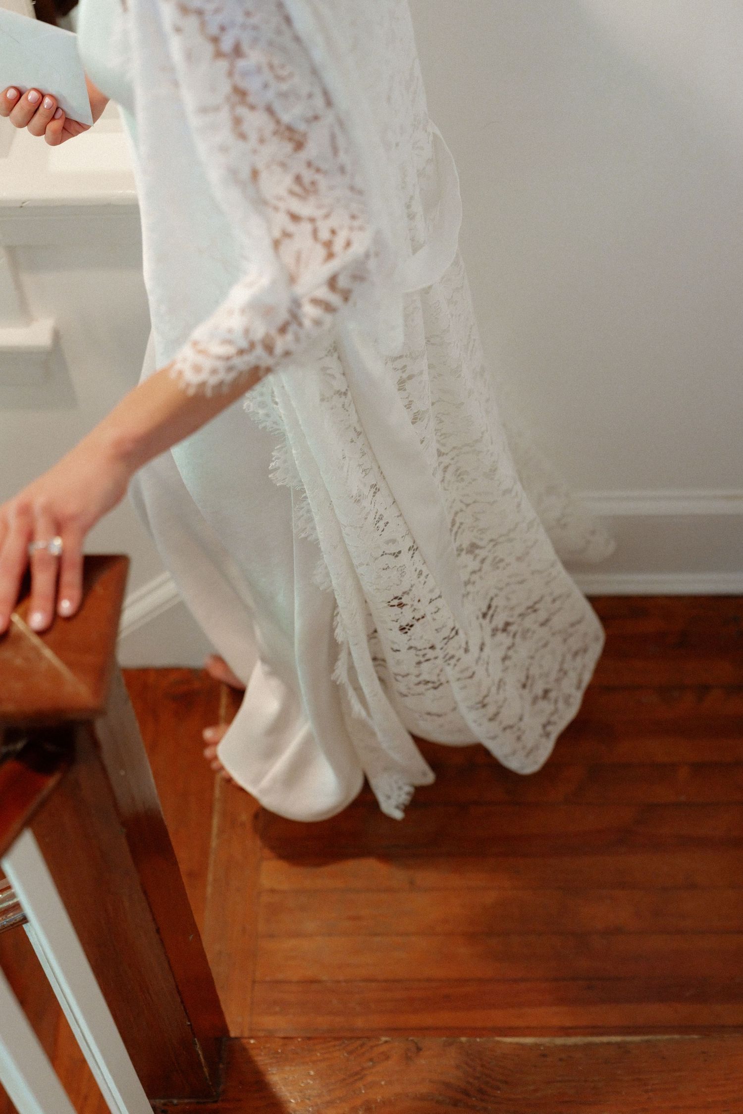 Bride in white lace wedding gown descending wooden staircase, diamond ring visible on hand.