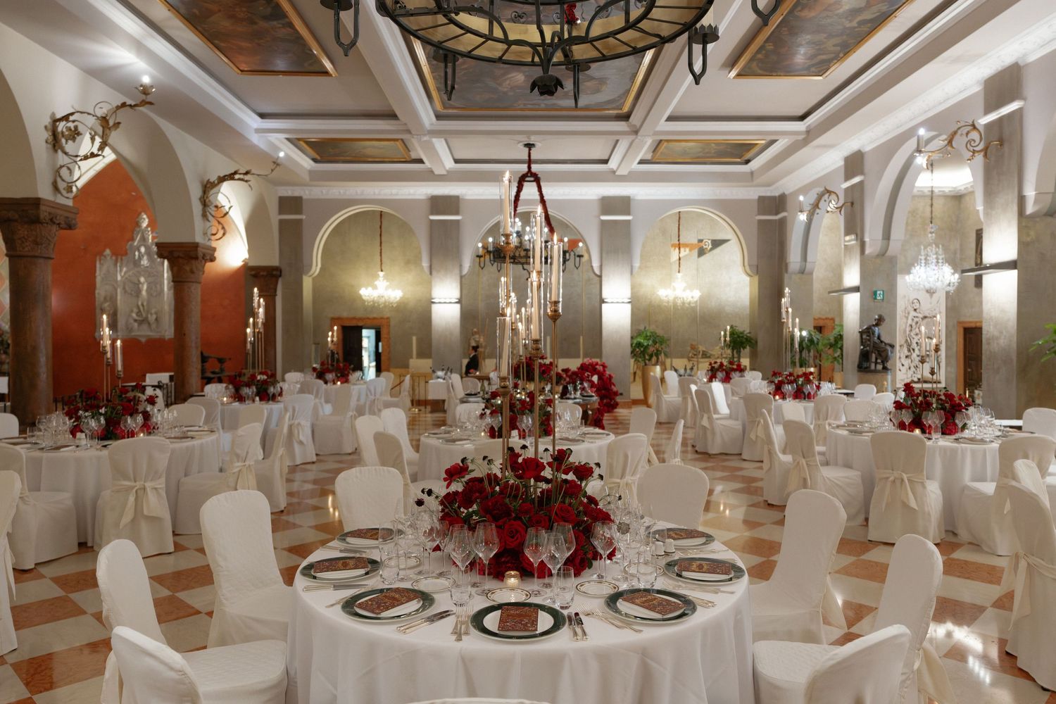 Elegant banquet hall with white-draped round tables, red floral centerpieces, chandeliers, and arched windows for weddings.