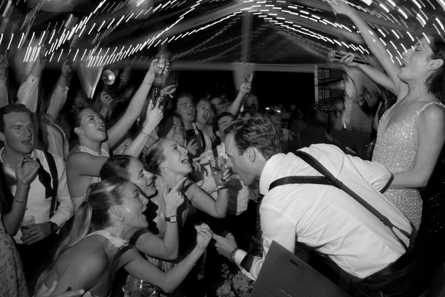 Black and white photo of a lively wedding reception dance floor with guests celebrating and raising their hands.