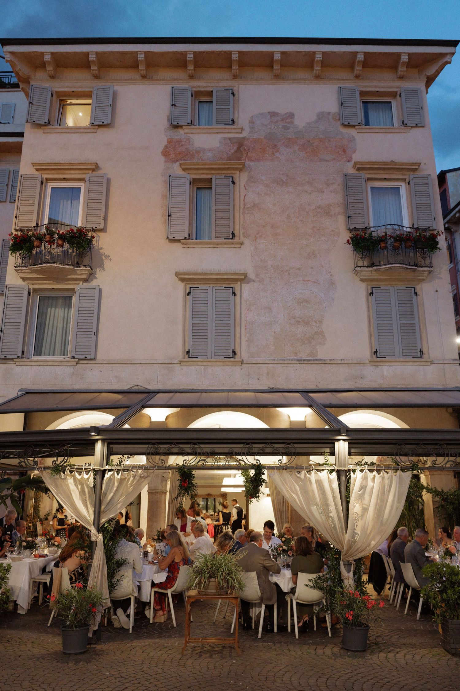 Outdoor restaurant terrace with diners seated under illuminated arched canopy against a classic Italian building facade at dusk.