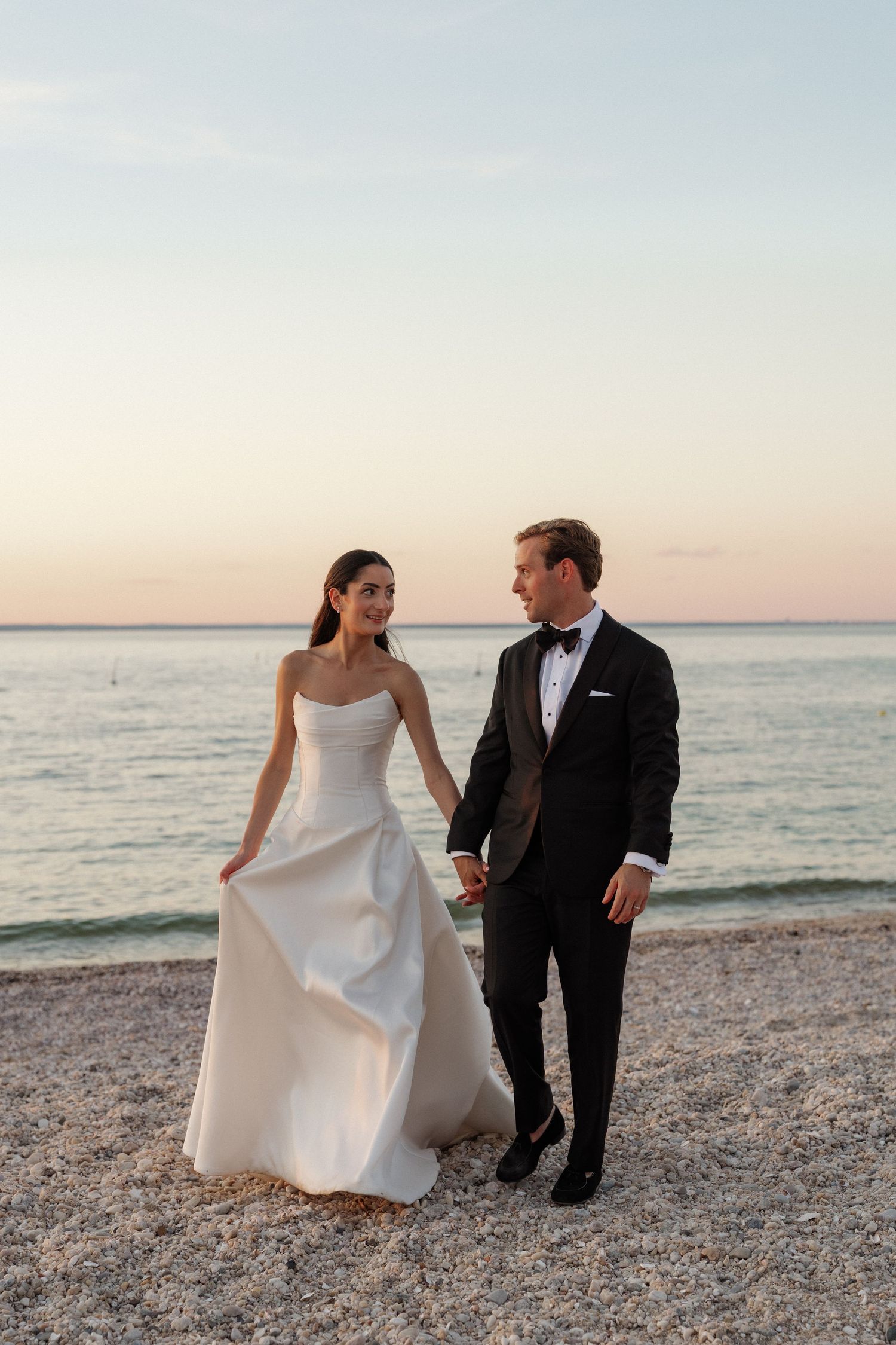 Bride and groom holding hands on a pebble beach at sunset, bride in white strapless gown, groom in black tuxedo.
