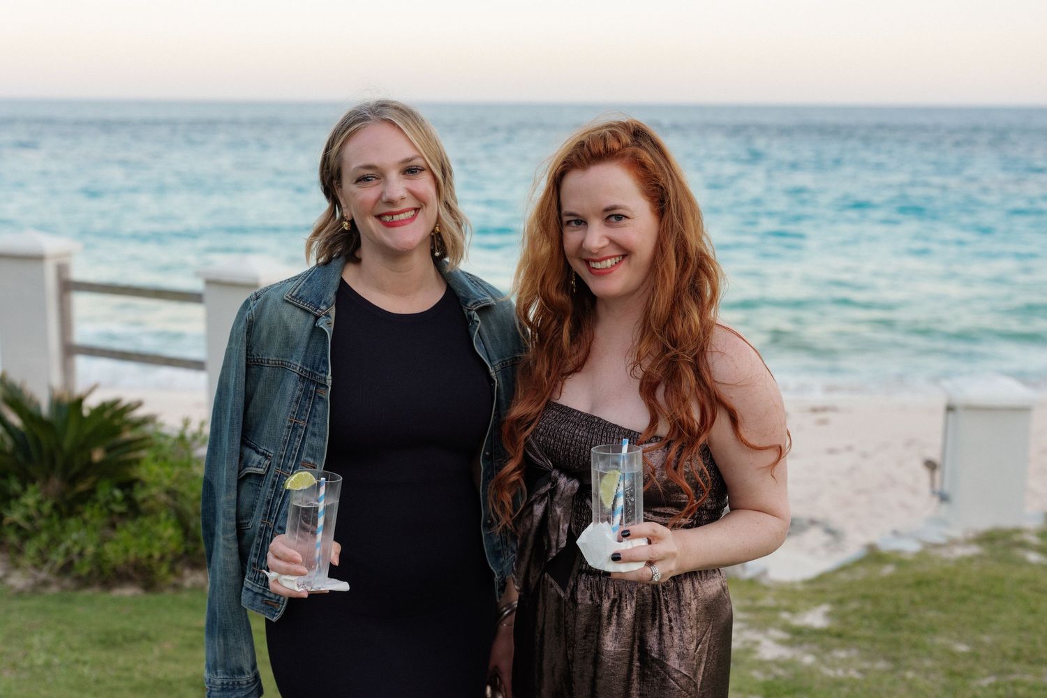 Two smiling women stand together on a beach with the ocean in the background.