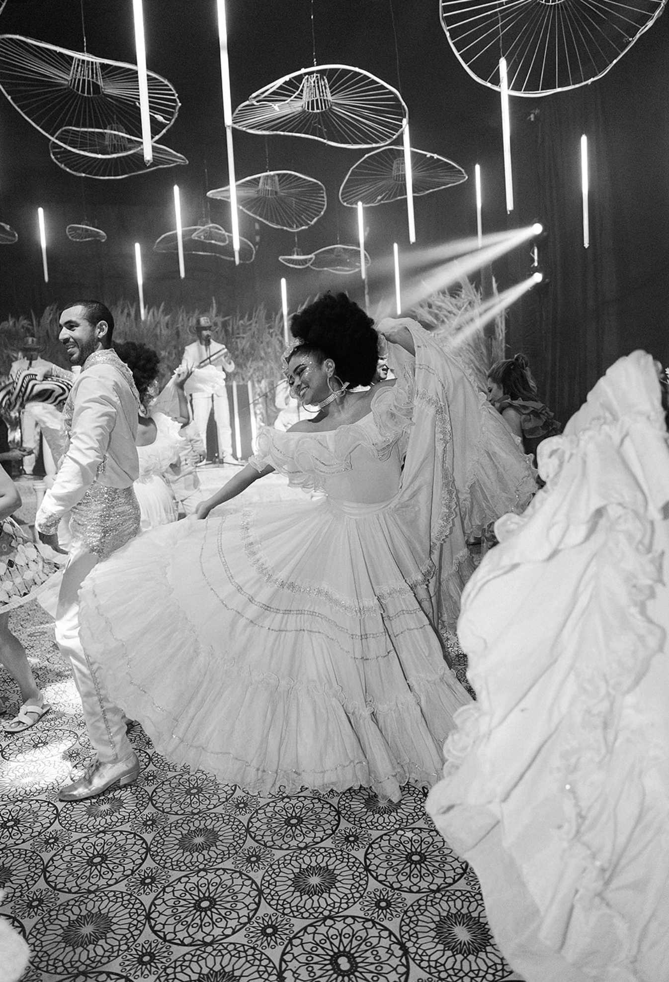 People in flowing white dresses twirl on a patterned dance floor under modern vertical lighting.