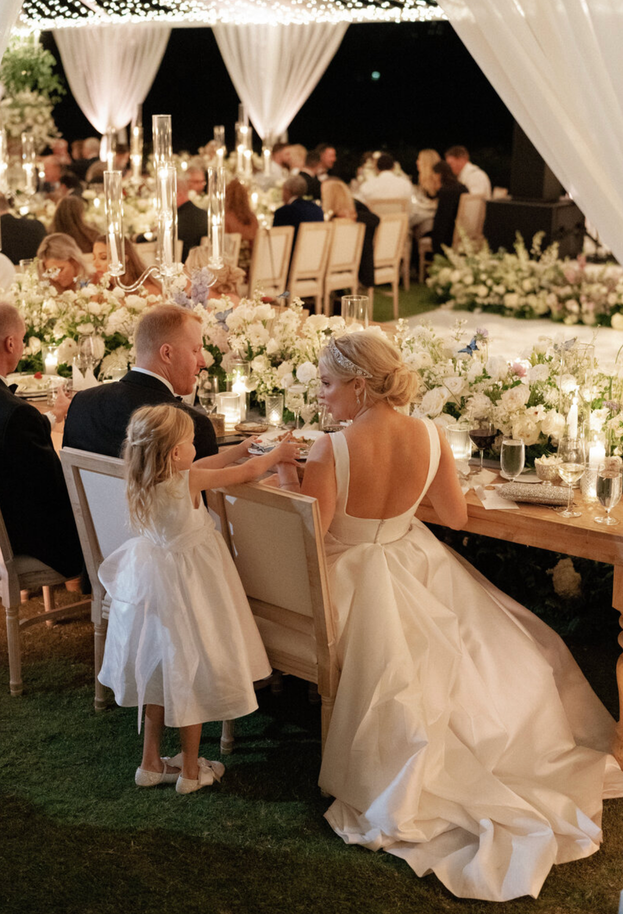 Wedding reception scene with elegant table settings, floral centerpieces and candlelight under draped fabric.