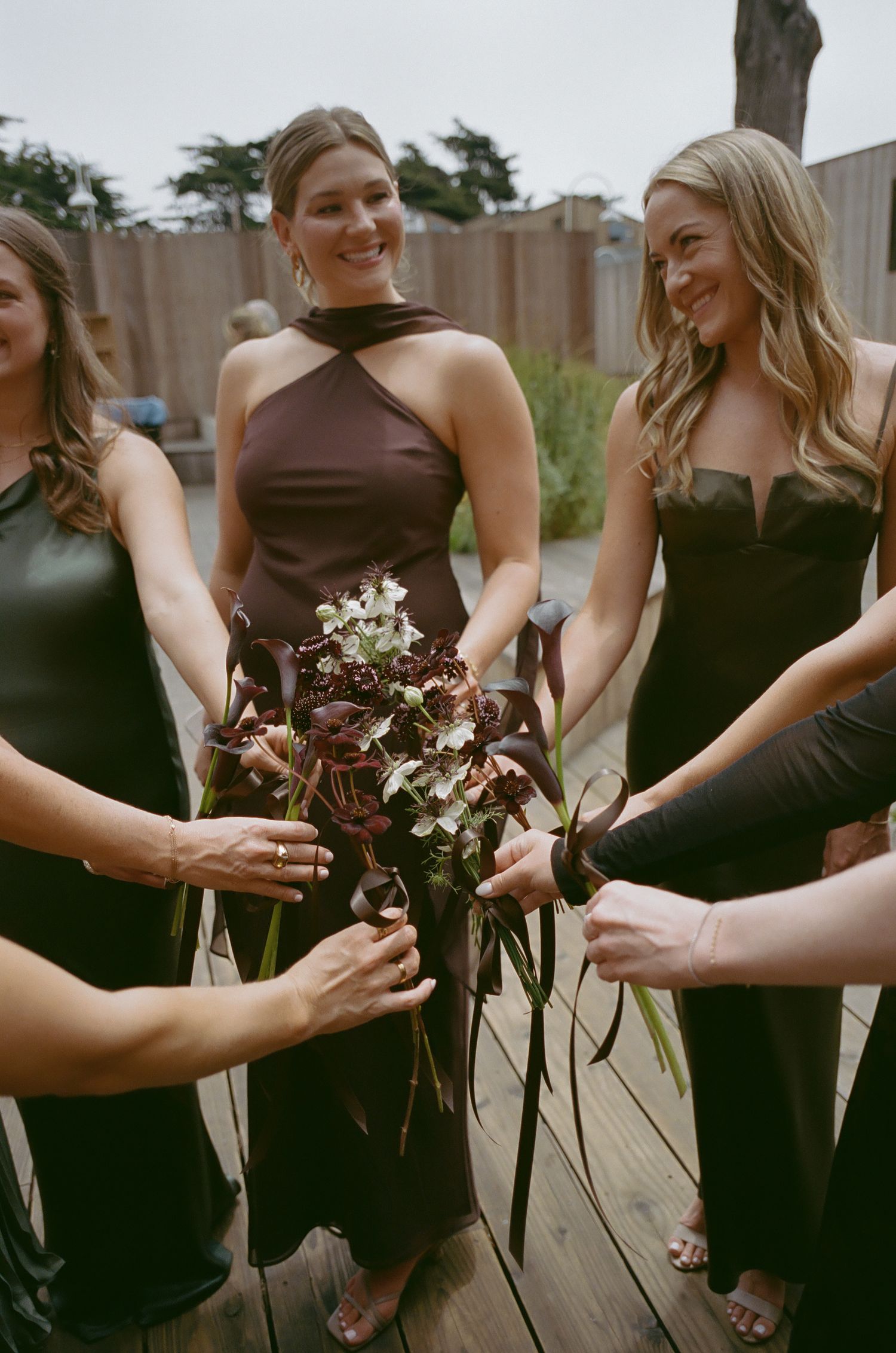 Bridesmaids in dark dresses hold burgundy and white floral bouquets together in an artistic arrangement.
