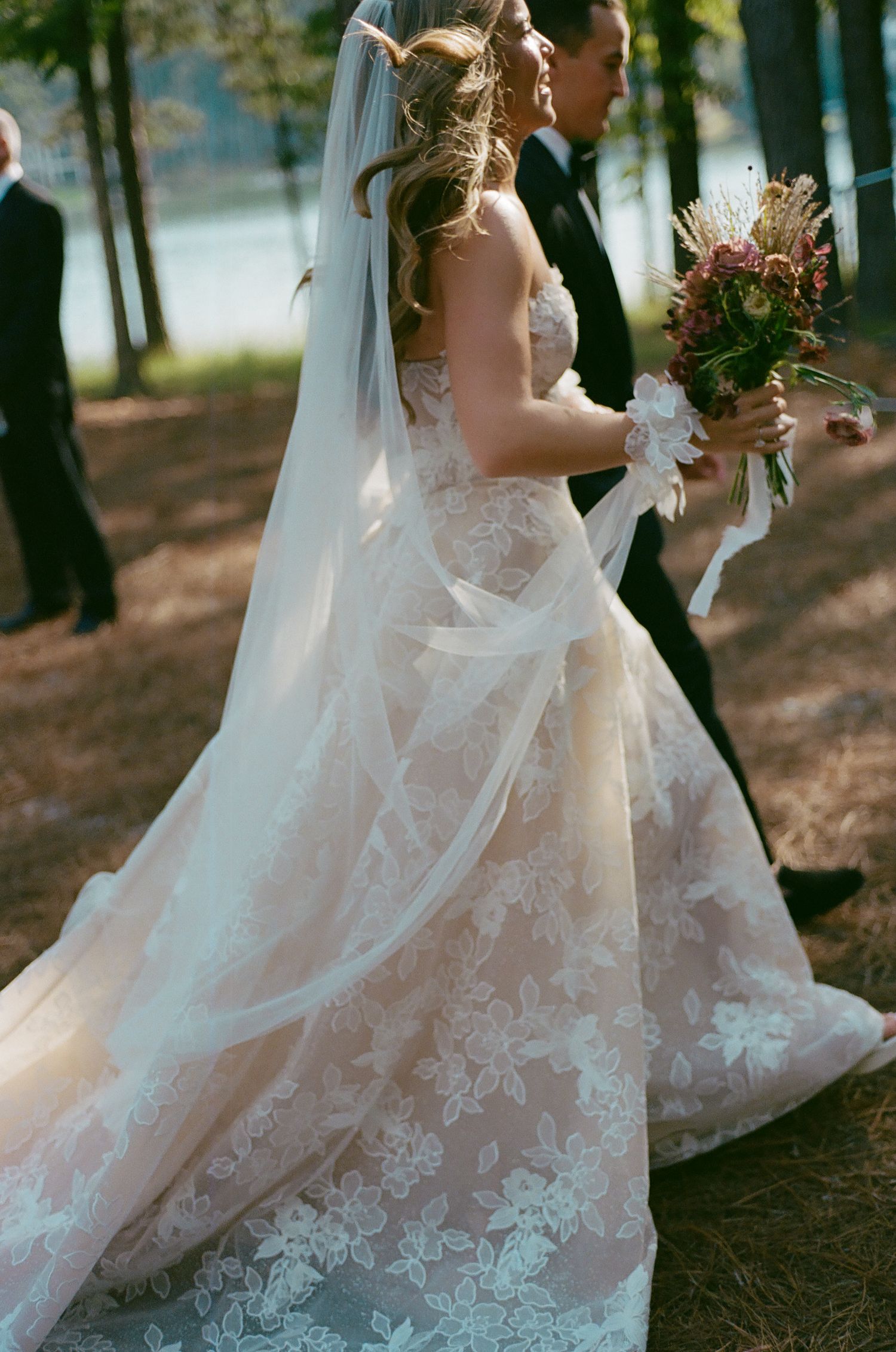Elegant lace wedding dress detail with flowing train captured in outdoor forest setting.
