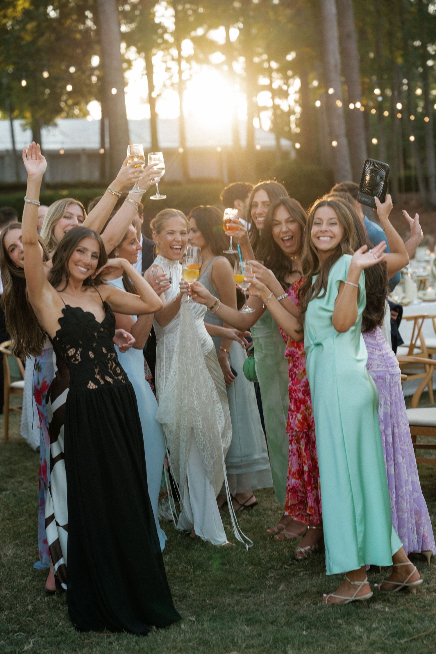 A celebratory group of wedding guests in colorful dresses toasting with champagne at sunset.