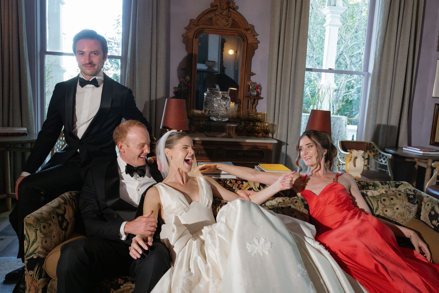 Elegant wedding party laughing together, bride in white gown, guests in formal attire in a luxurious room.