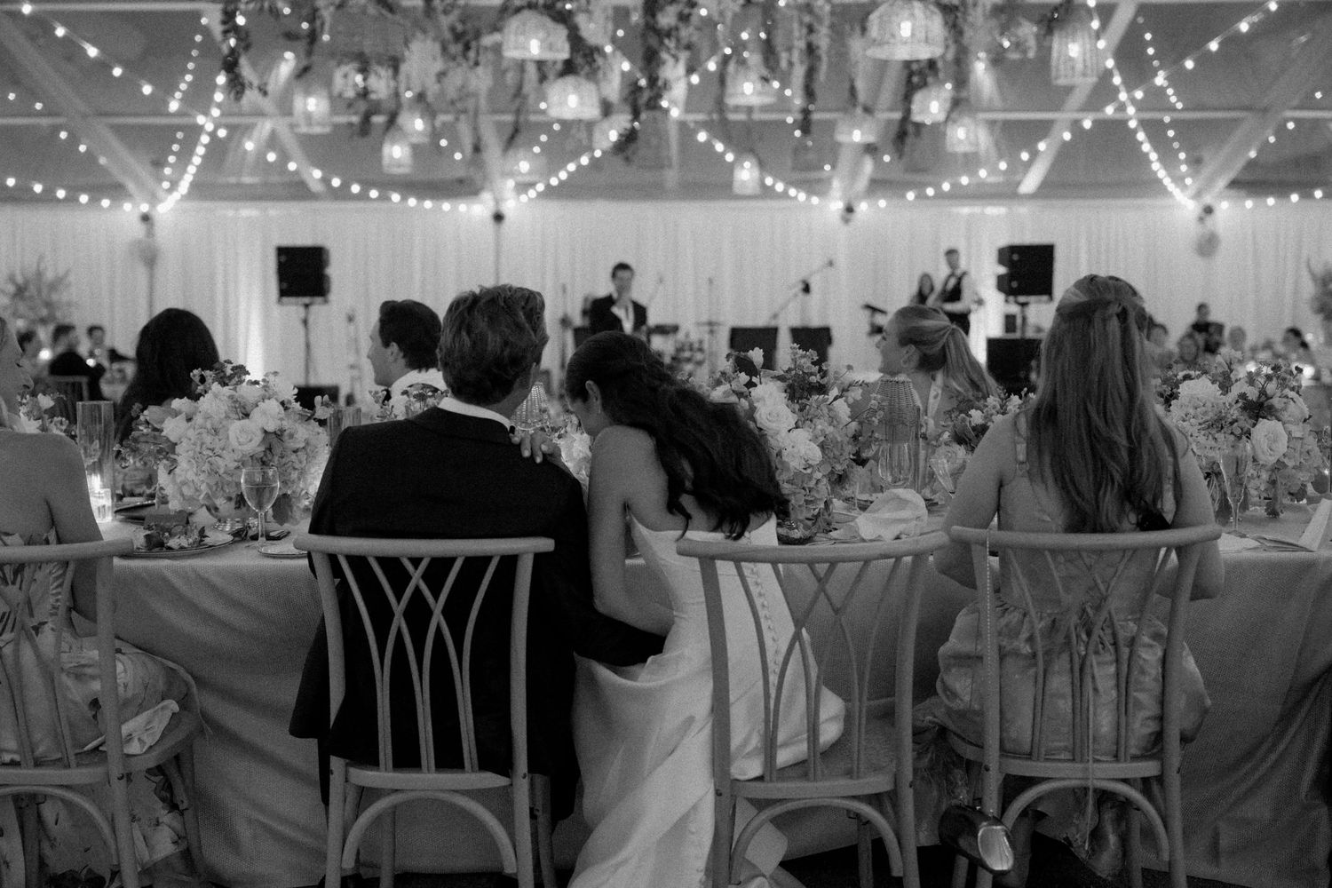 Black and white wedding reception photo of couple embracing at table under string lights.
