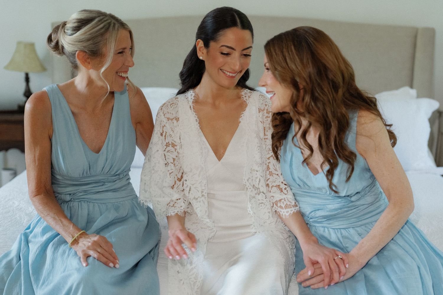Bride in lace wedding dress sitting on bed smiling with two bridesmaids in light blue gowns.