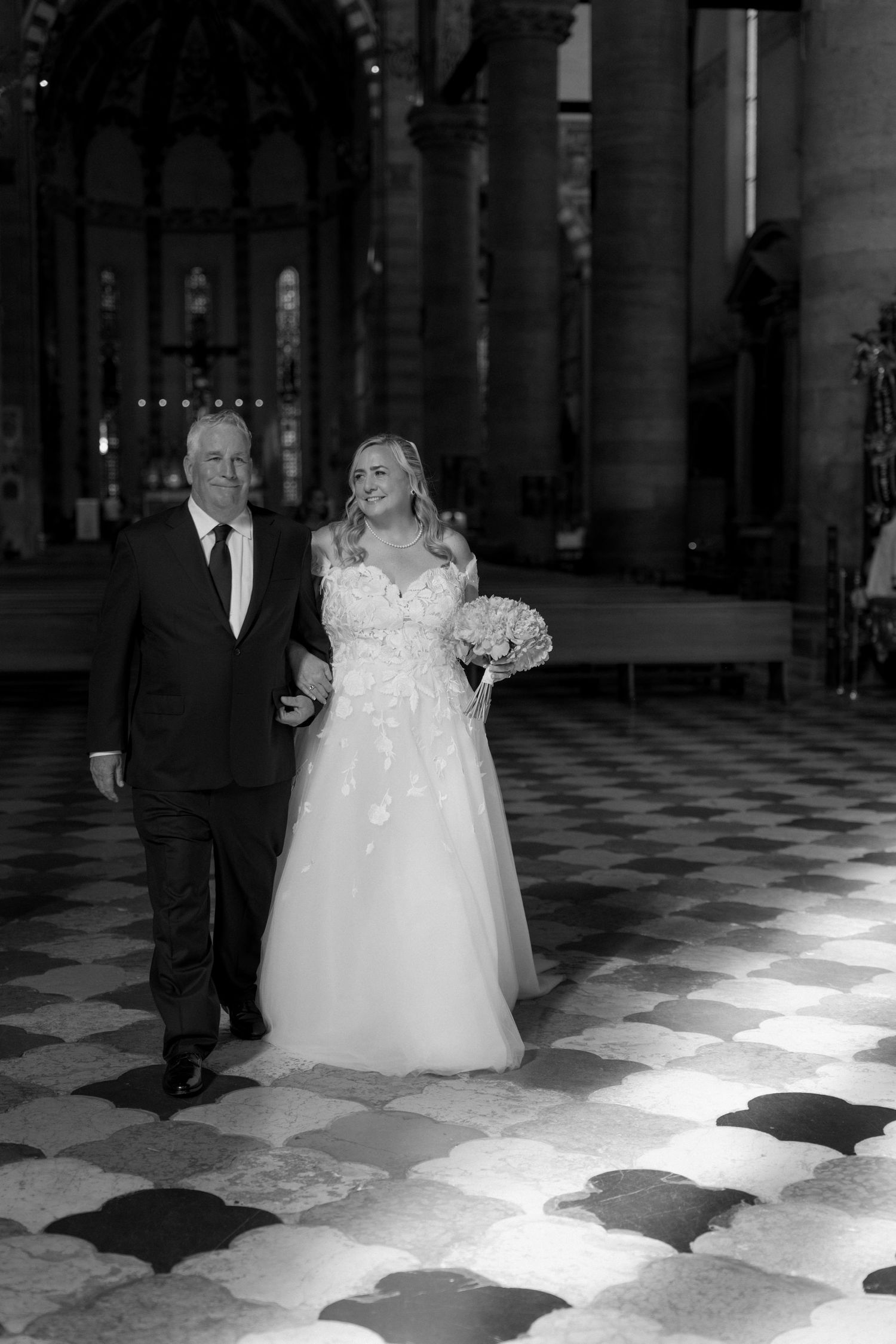 Bride in white gown walks down church aisle with escort on checkered floor in elegant black and white wedding photo.
