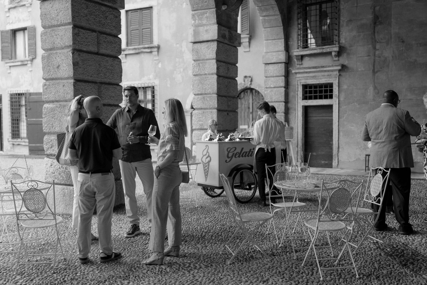 Black and white photo of people gathered near a gelato cart under stone archways in an Italian piazza.