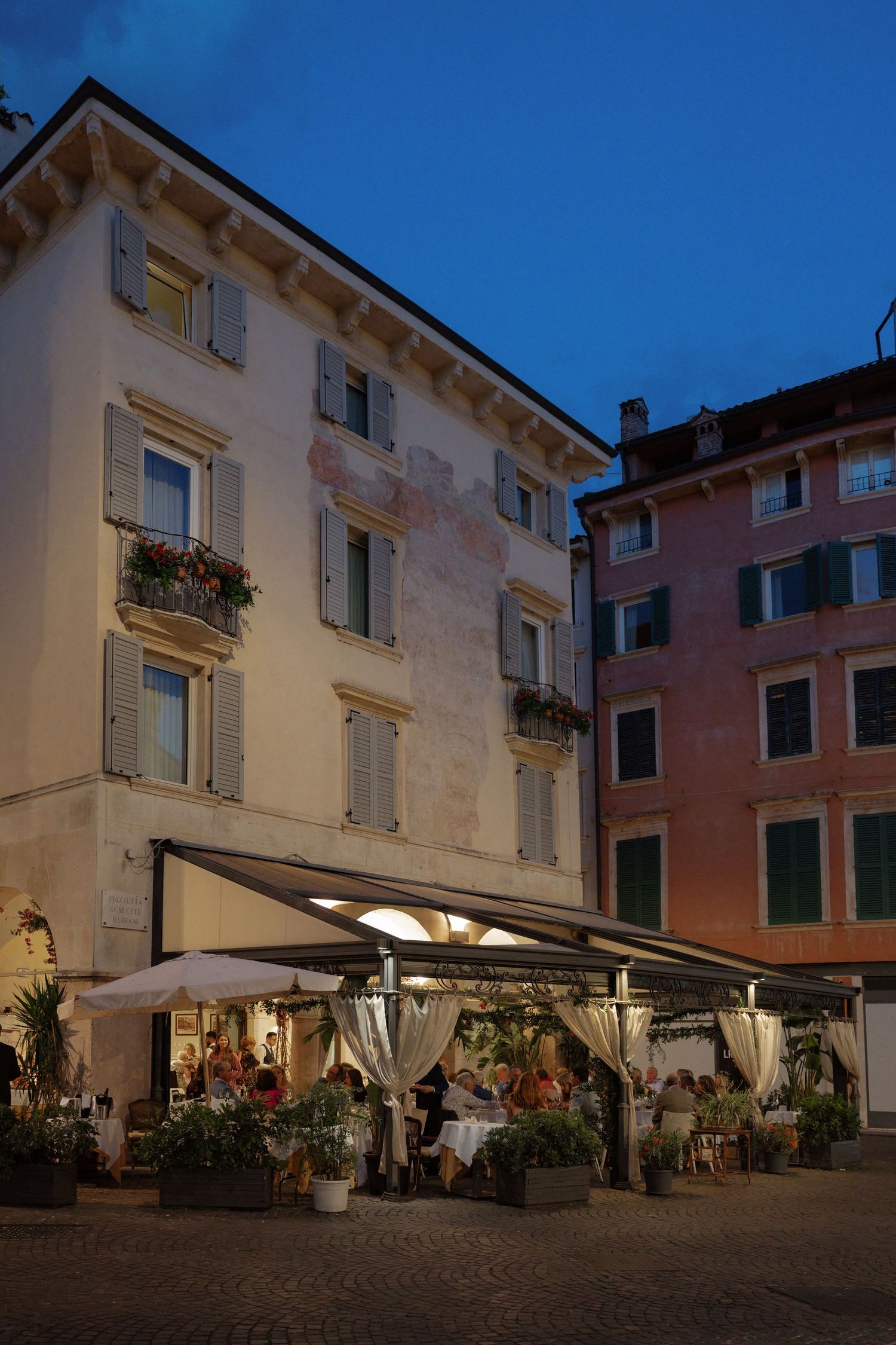 Outdoor restaurant terrace lit at dusk beneath historic Italian buildings in a charming cobblestone piazza.