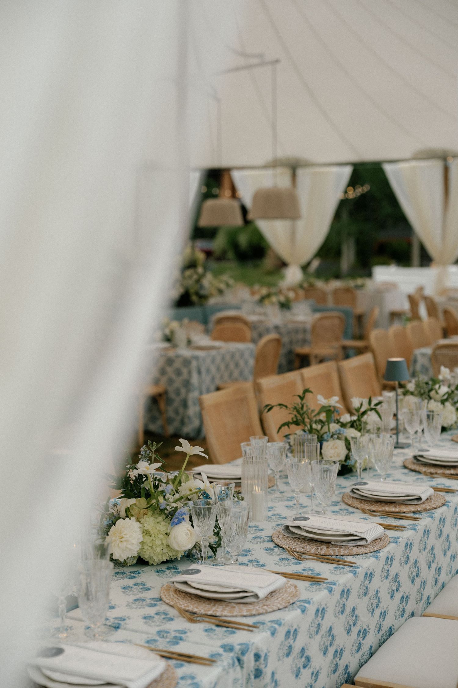 Long dining tables with blue patterned tablecloths and white place settings under a white tent canopy.