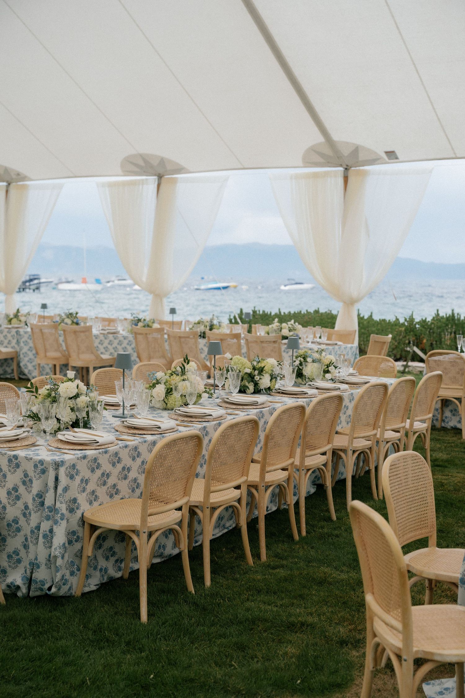 Elegant outdoor wedding reception setup with white tent, floral tablecloths and rattan chairs overlooking scenic views.