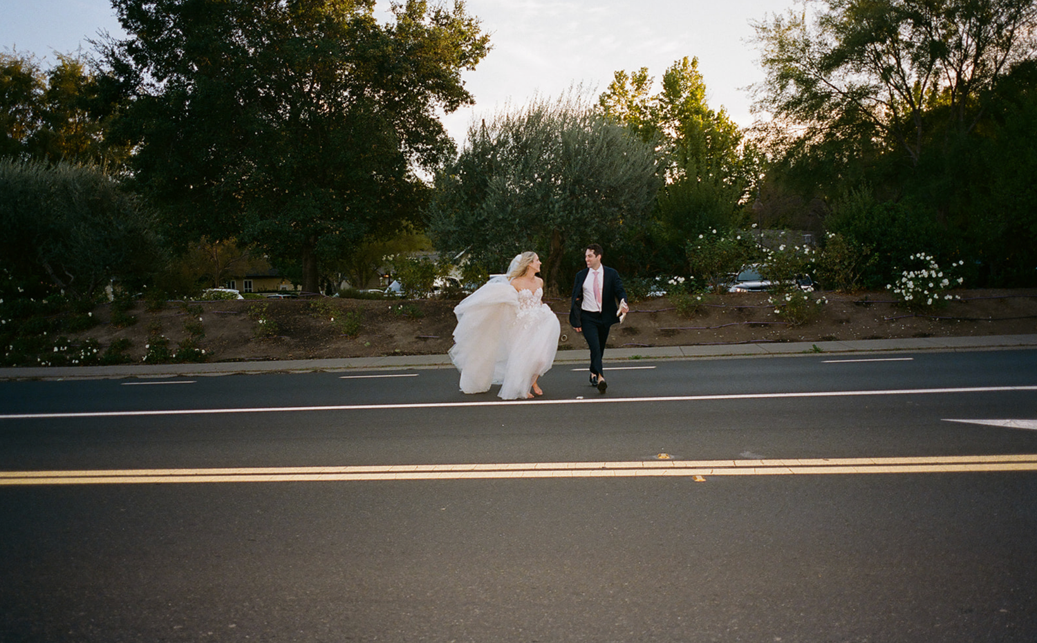 A couple walking on a road during sunset, with the bride's dress flowing in the breeze.