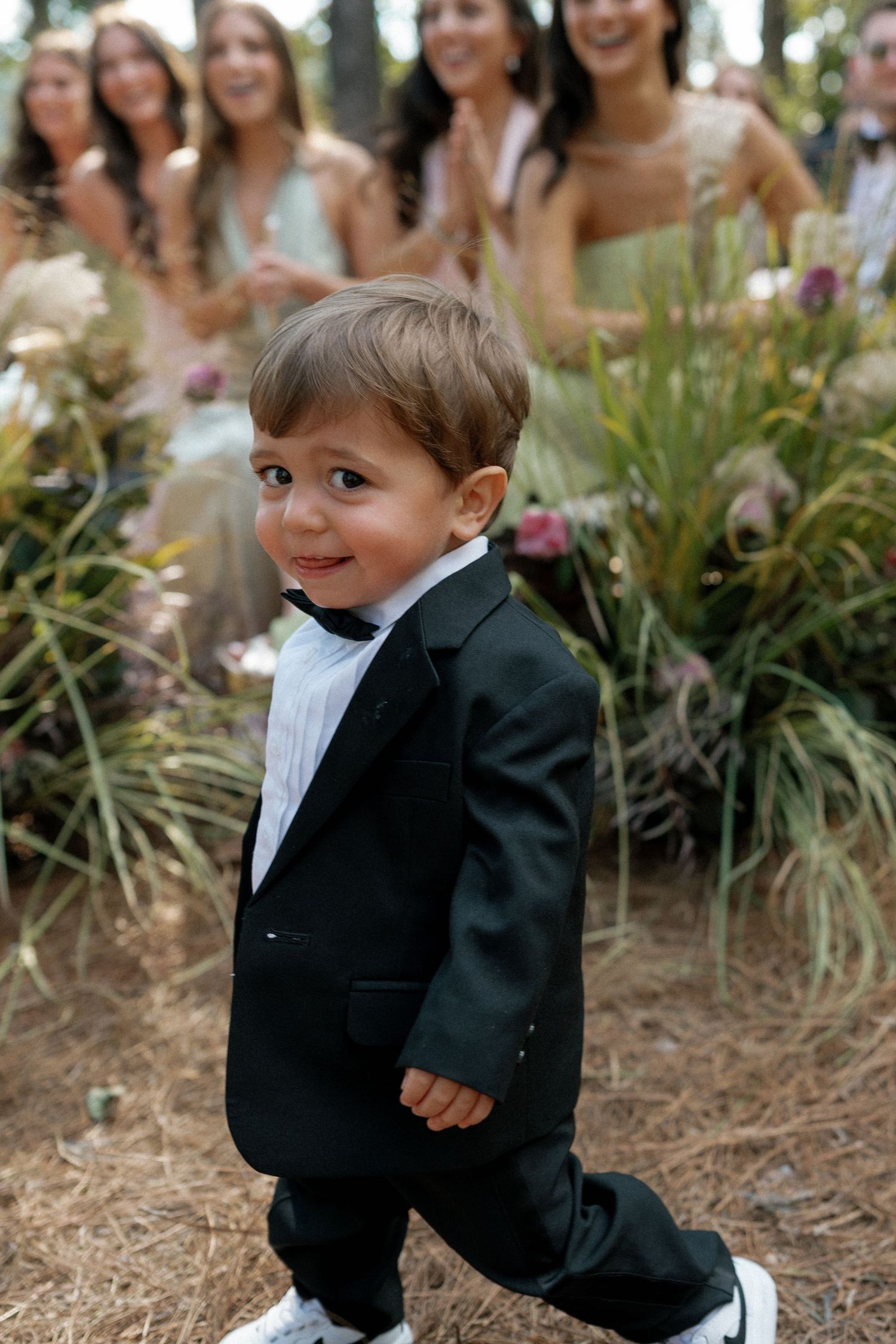 Young ring bearer in a black tuxedo walking down the aisle at an outdoor wedding.