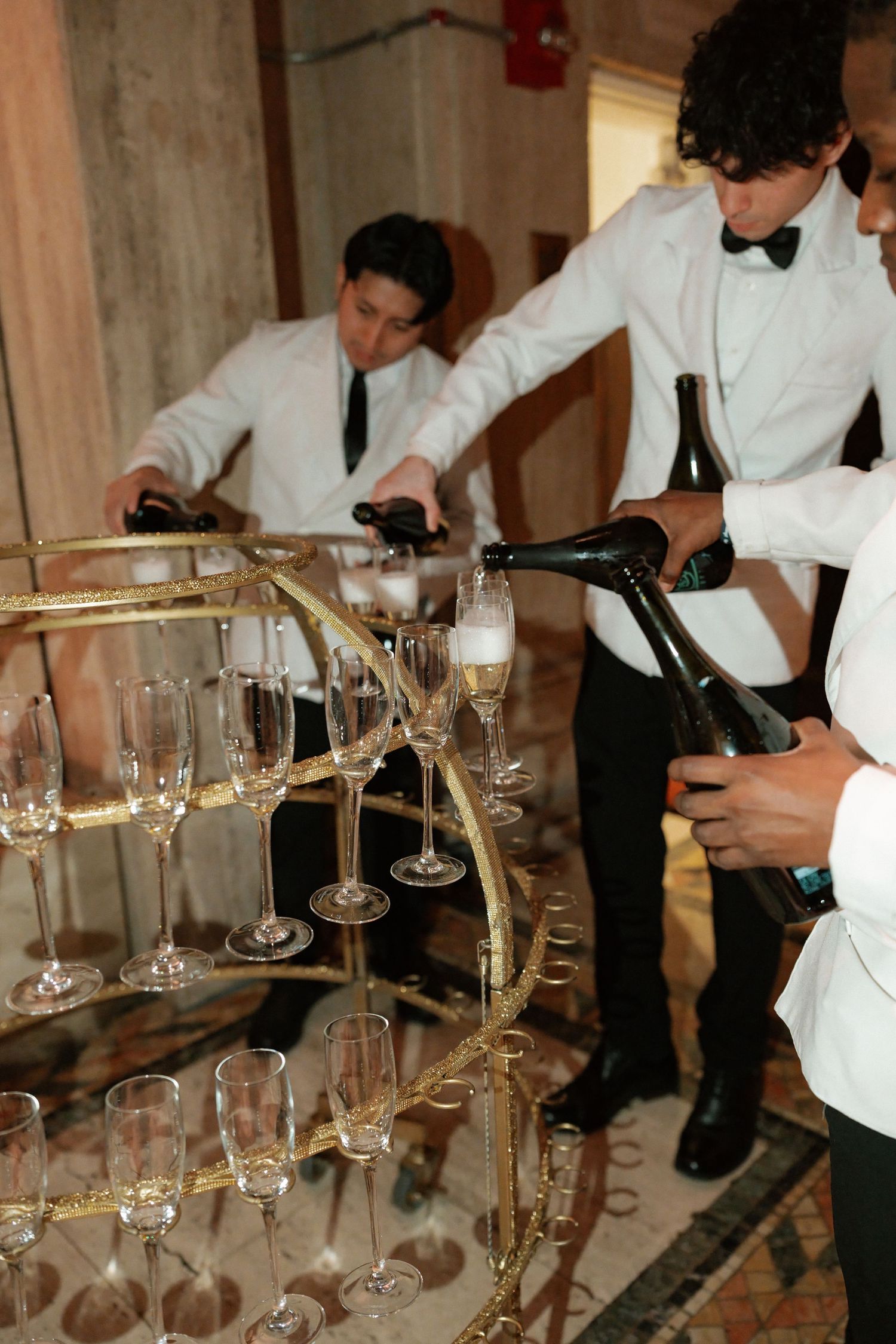 The image depicts two men in formal attire pouring champagne into glasses on a table set for a formal event.