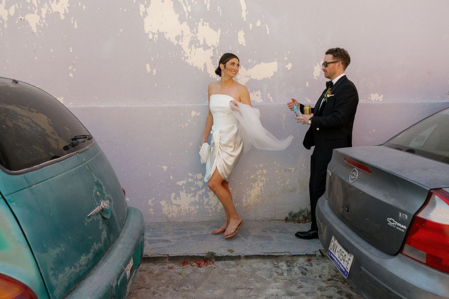 Newlyweds share a playful moment between parked cars on their wedding day.
