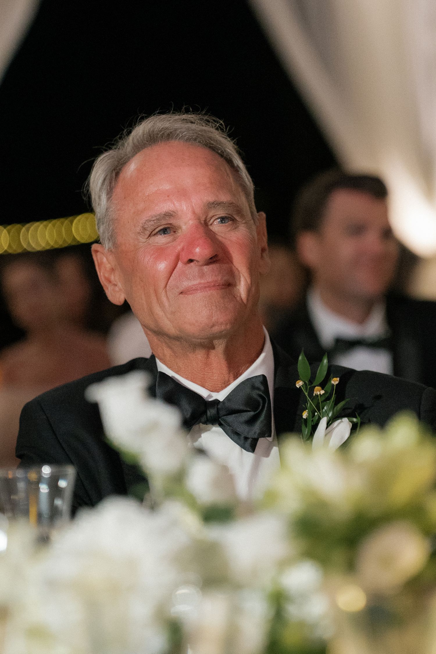 A guest at a formal wedding reception wearing a black bow tie smiles warmly.