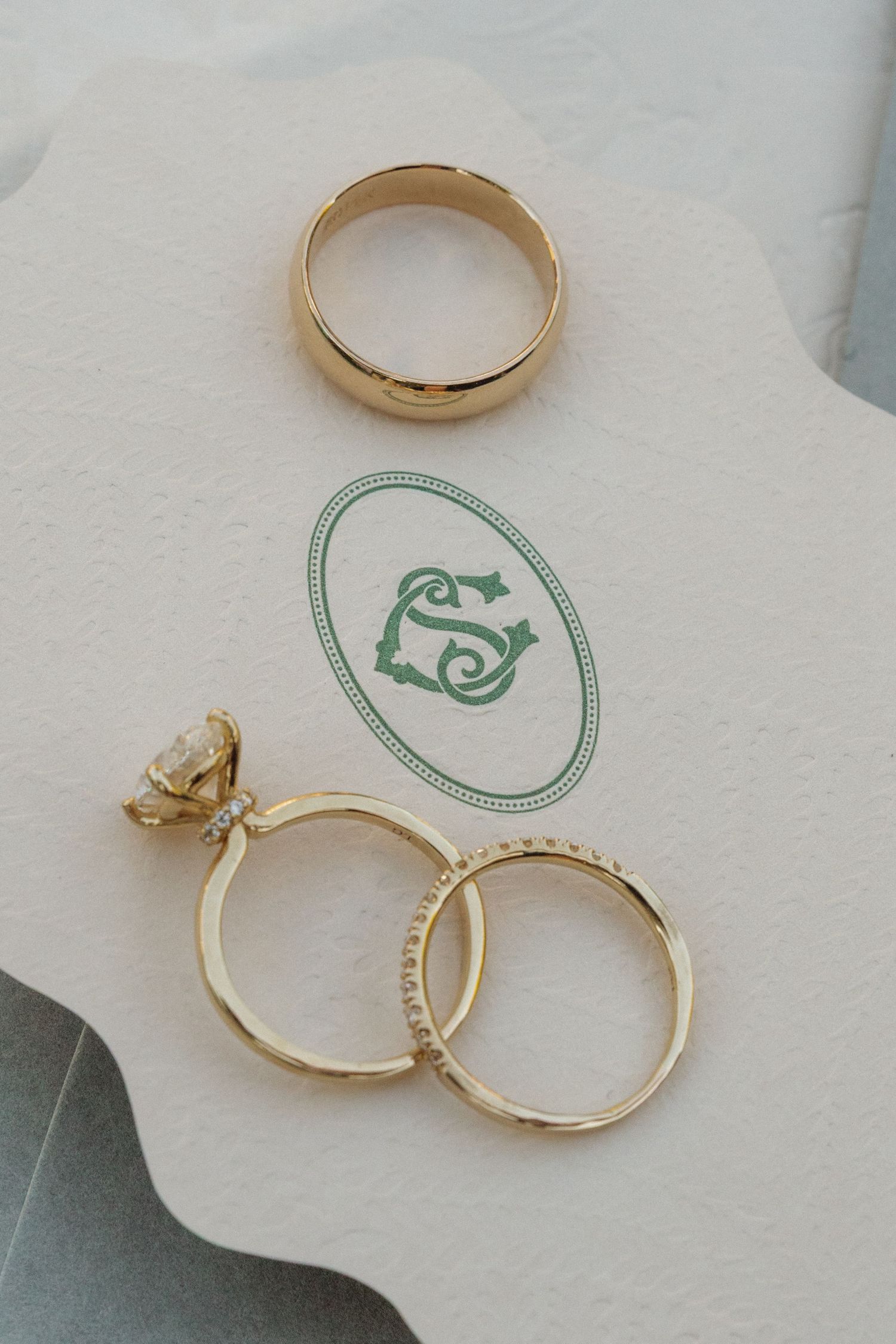 Gold wedding rings and engagement ring displayed on white paper with a green monogram logo.