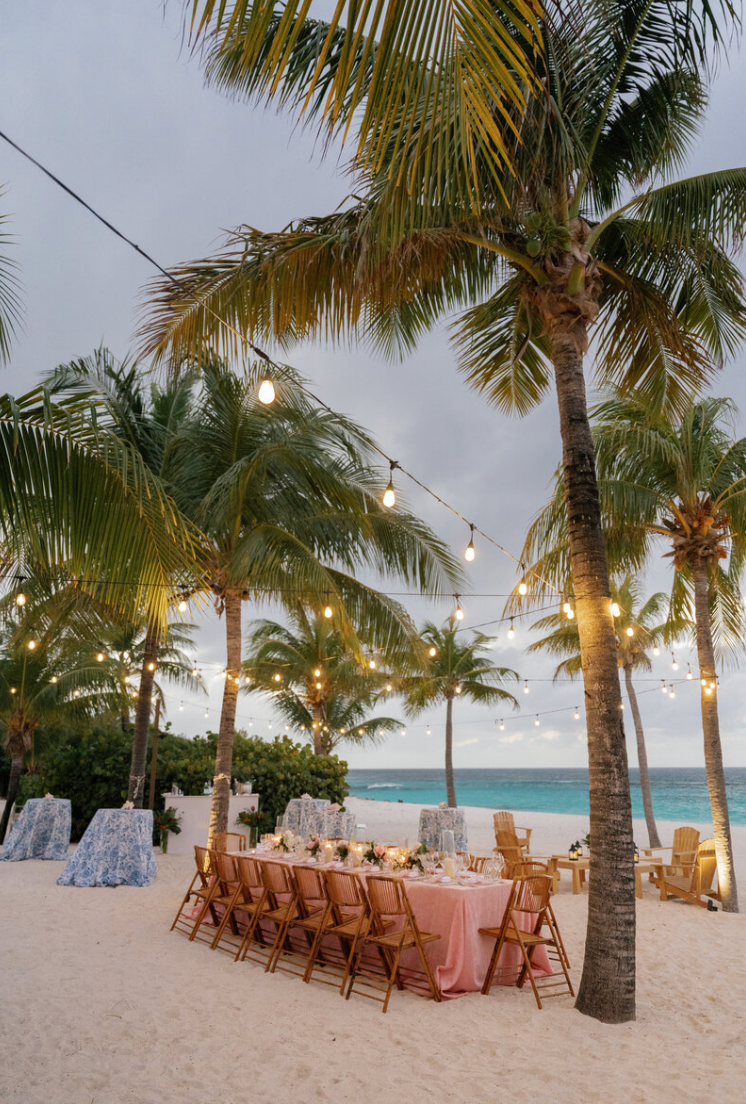 Tropical beach wedding setup with wooden chairs and palm trees against turquoise ocean backdrop.