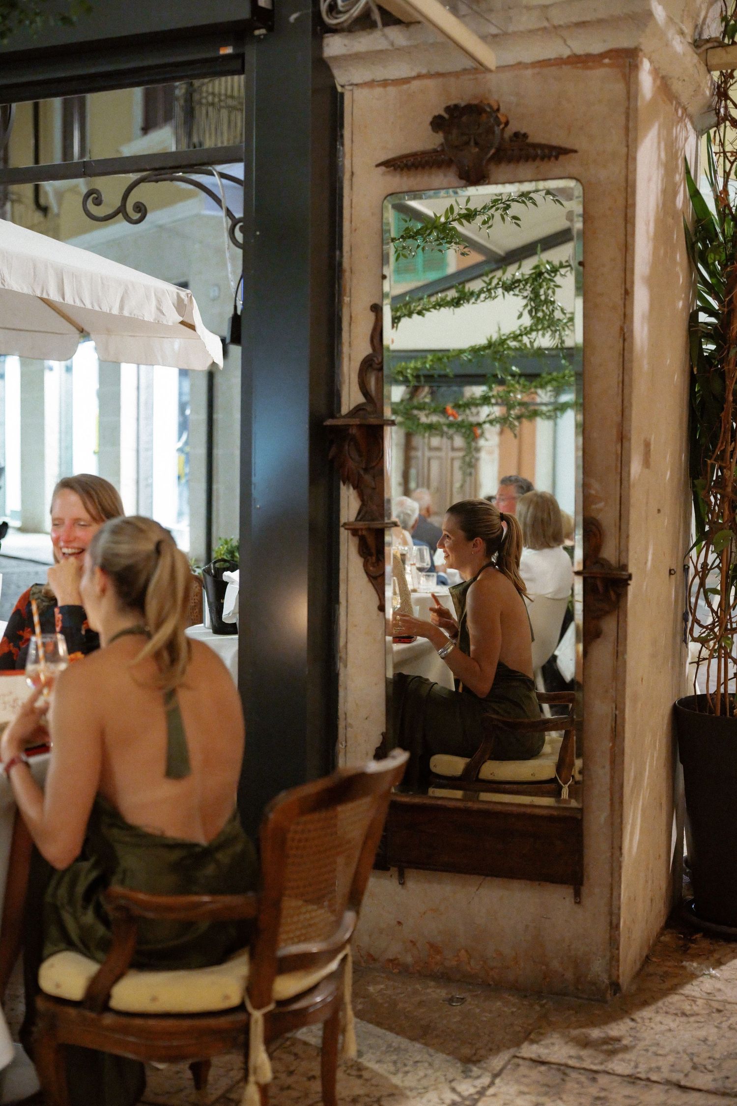 Two women in elegant dresses dining at a rustic Italian restaurant, reflected in an ornate vintage mirror.