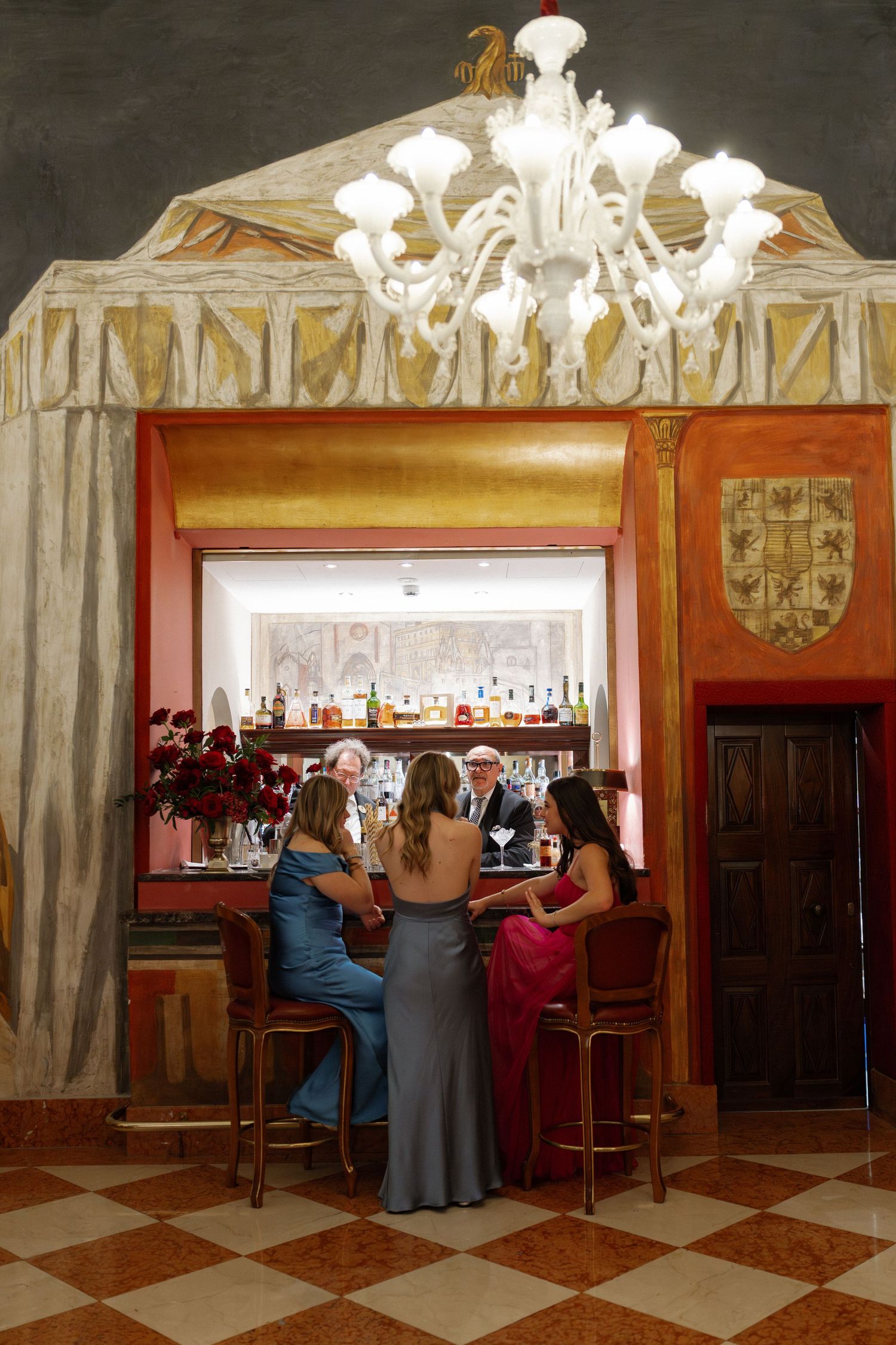 Three elegantly dressed women socializing at a ornate bar with Murano glass chandelier overhead.