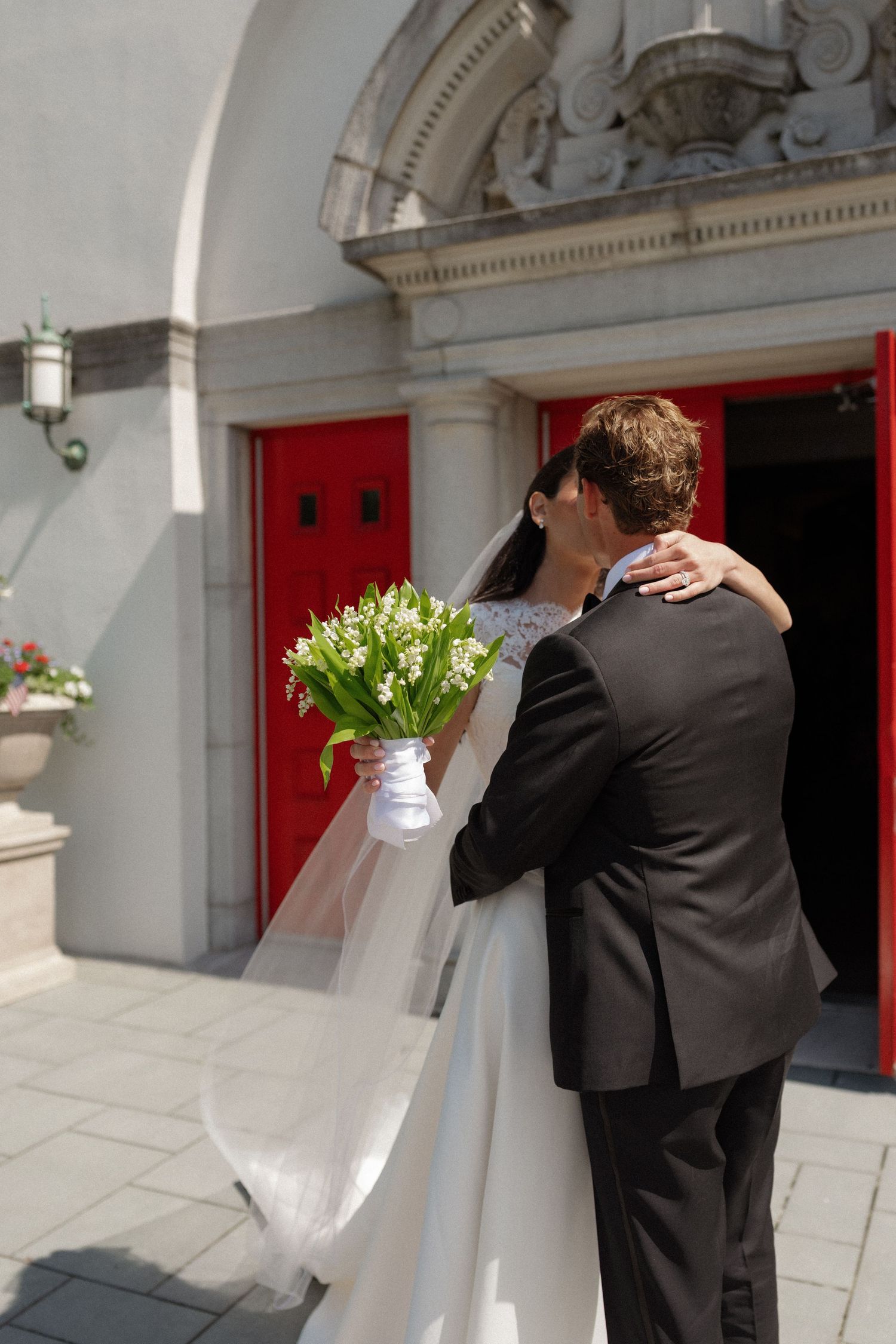 Bride and groom share a kiss outside a church with red doors, bride holding a white lily of the valley bouquet.