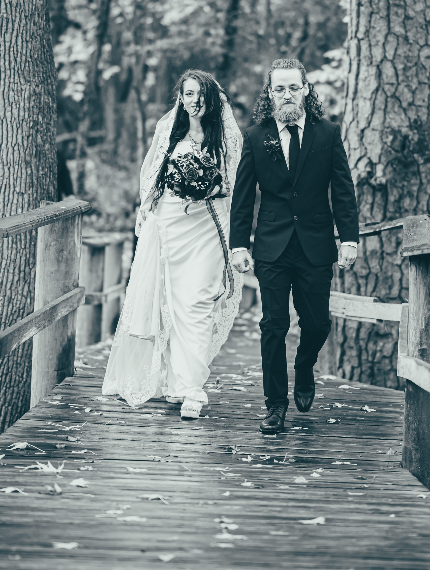Black and white photo of a couple walking down a wooden path through trees.